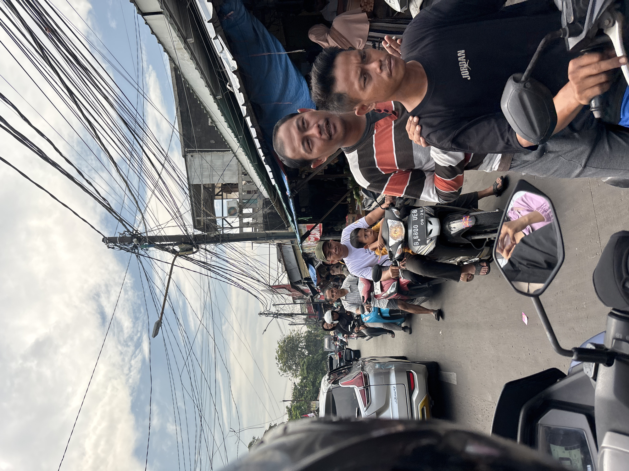 The image depicts a bustling street scene in North Jakarta, Indonesia, observed from the perspective of a scooter rider. Overhead, a dense web of electrical wires stretches across the sky. The street is congested with motorbikes and a single silver car. Several people are riding motorbikes, with some carrying passengers. Two men on a motorcycle at the top of the frame are looking back and smiling at the camera. The mirror shows another passenger on a motorcycle behind them. The scene is indicative of urban life in Jakarta.