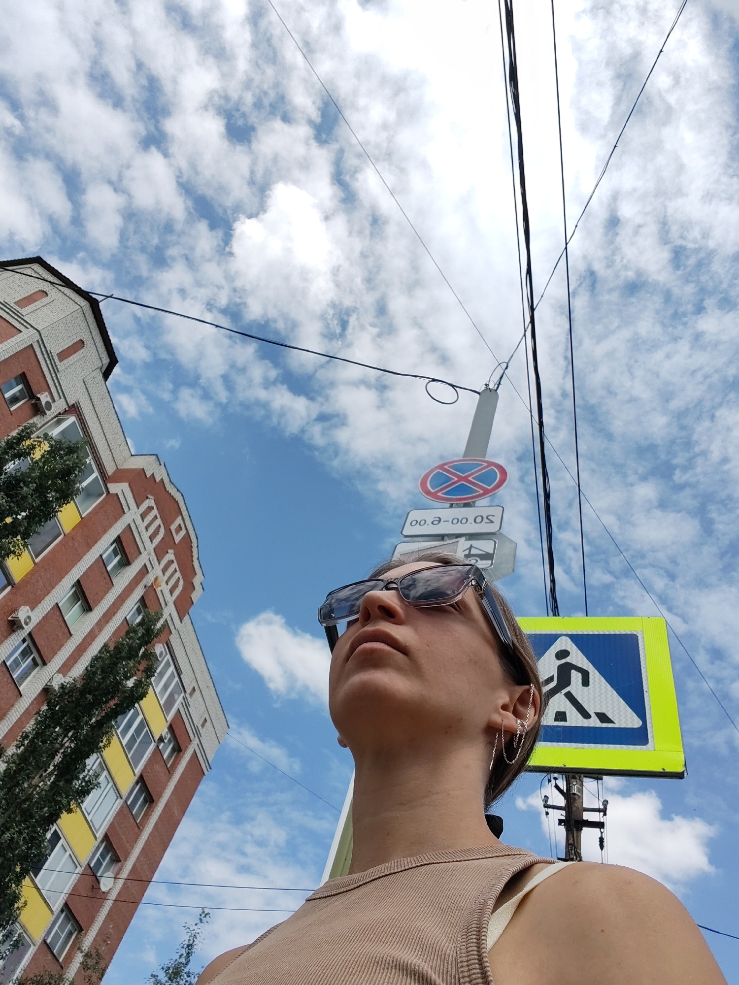 A woman wearing sunglasses and a beige tank top is standing outside, looking upwards. She has an ear cuff on her left ear. Behind her, there is a multi-story brick building with white and yellow accents. A street signpost with multiple signs, including a "no stopping" sign, a sign with Cyrillic text, a sign with a bridge graphic, and a pedestrian crossing sign, is visible. Overhead power lines crisscross the bright blue sky filled with scattered white clouds. The perspective is from a low angle, looking up at the woman and the surrounding environment.