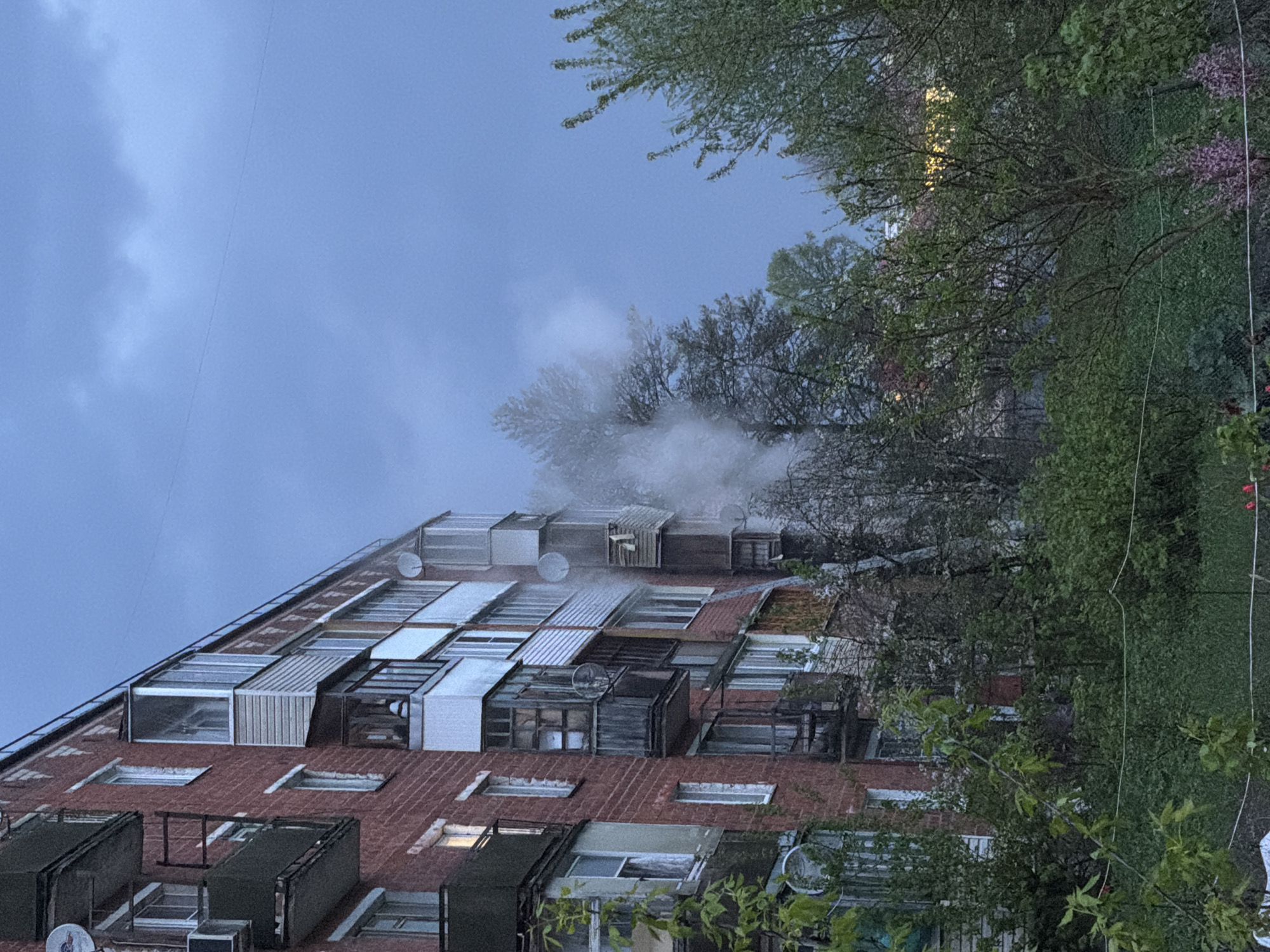 The image shows a multi-story brick building with numerous windows, some of which appear to be enclosed or covered. A substantial amount of smoke or steam is rising from the building, possibly from the top floors. The sky is overcast. Trees with green foliage are visible to the right of the building. The location is suggested to be Reshetnikovo, Russia.