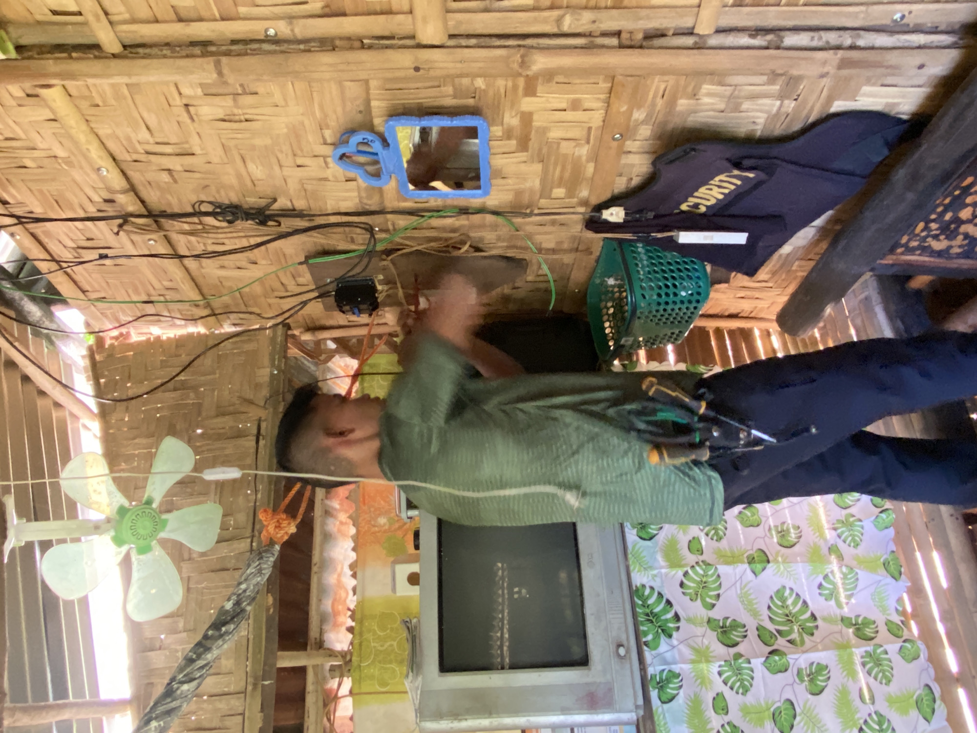 The image depicts a man working on electrical wiring inside a structure with walls made of woven bamboo. He is wearing a green striped t-shirt and dark pants, and has a tool belt with several tools around his waist. To his left is an old television set on a shelf covered with a patterned fabric. Above him, a white and green oscillating fan hangs from the ceiling. To his right, various electrical wires are visible, some connected to a black electrical box. A green plastic basket hangs on the wall, and below it is a dark blue vest with the word "SECURITY" in yellow lettering. A small blue-framed mirror hangs on the wall to the right of the electrical wiring. The overall environment suggests a humble dwelling or workshop, possibly in a rural or less developed area of Davao City, Philippines, given the materials used for construction and the style of the appliances.