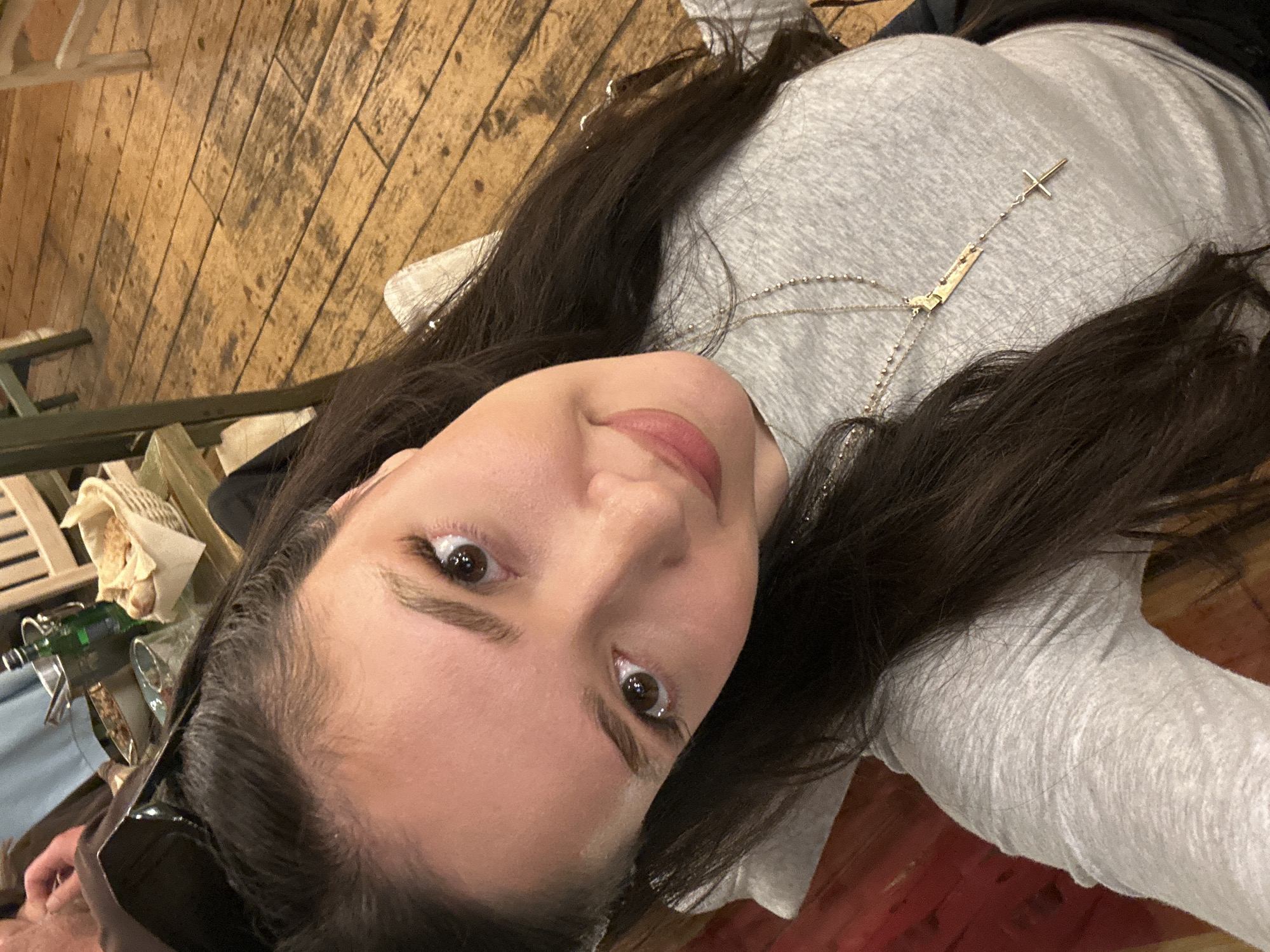 A woman with brown hair is shown from a low, canted angle, looking upward at the camera. She wears a gray, long-sleeved shirt and a delicate necklace with a cross pendant. Sunglasses are perched on her head. The background includes a wooden table, chair, and a woven basket containing bread. A partially visible bottle and glass are also present. The location appears to be Tsakhkadzor, Armenia.