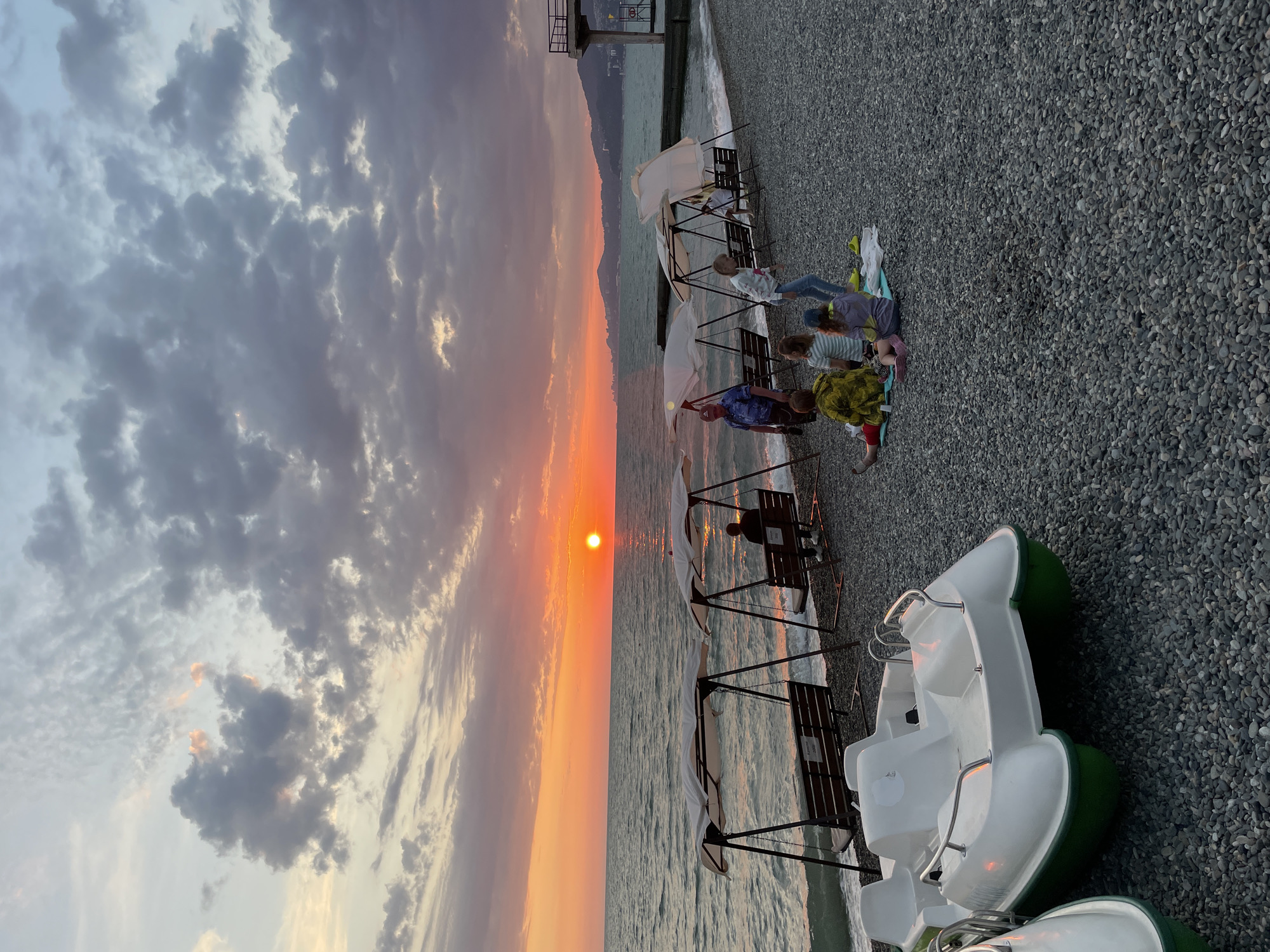 This is a vertical, eye-level, full-shot photograph of a beach in Sochi, Russia, during sunset. The sky is filled with dramatic clouds, ranging from soft pinks and oranges near the horizon to darker grays and purples overhead. The sun, a bright orange orb, is setting over the Black Sea, casting a shimmering reflection on the water.
On the pebble beach, several people are gathered. In the foreground, two white and green pedal boats are parked. Further back, a group of children and adults are sitting on a blanket, seemingly having a picnic. A few swings with benches are set up along the shoreline, with one person seated on a swing. The sea gently laps at the shore, and in the distance, mountains are visible under the twilight sky. A concrete pier extends into the sea on the right side of the image. The overall atmosphere is peaceful and serene, capturing the beauty of a sunset on the coast.
