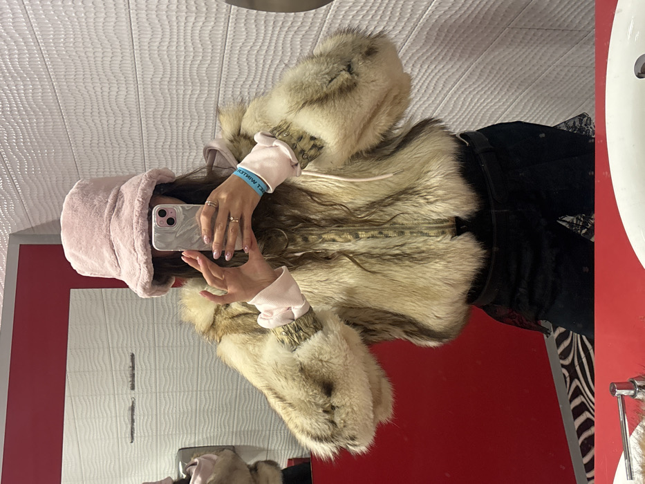 An individual is depicted taking a mirror selfie in a modern indoor space, likely a restroom or changing area. The person wears a light pink faux fur bucket hat and a multi-toned natural fur jacket with prominent sleeves, featuring light beige, brown, and darker streaked patterns, zipped up the front. Beneath the jacket, a light pink long-sleeved garment with ruffled cuffs is visible. Dark-colored bottoms, possibly shorts or a skirt with a black lace overlay and a black belt, are worn. The individual's right hand holds a light-colored smartphone with a prominent camera array, positioned to capture the reflection. Both hands display light pink manicured fingernails and multiple gold-colored rings. A blue wristband with reversed white text is present on the right wrist. The background, reflected in the mirror, consists of a solid red wall section on the left and a white textured wall with a wavy pattern on the right. In the unreflected foreground, a red counter spans the bottom, featuring a white porcelain sink basin with a chrome faucet on the right. A black and white zebra-patterned floor mat is partially visible in the lower-left corner. The specific location within the city of Krasnoyarsk, Russia, cannot be determined from the visual information provided.