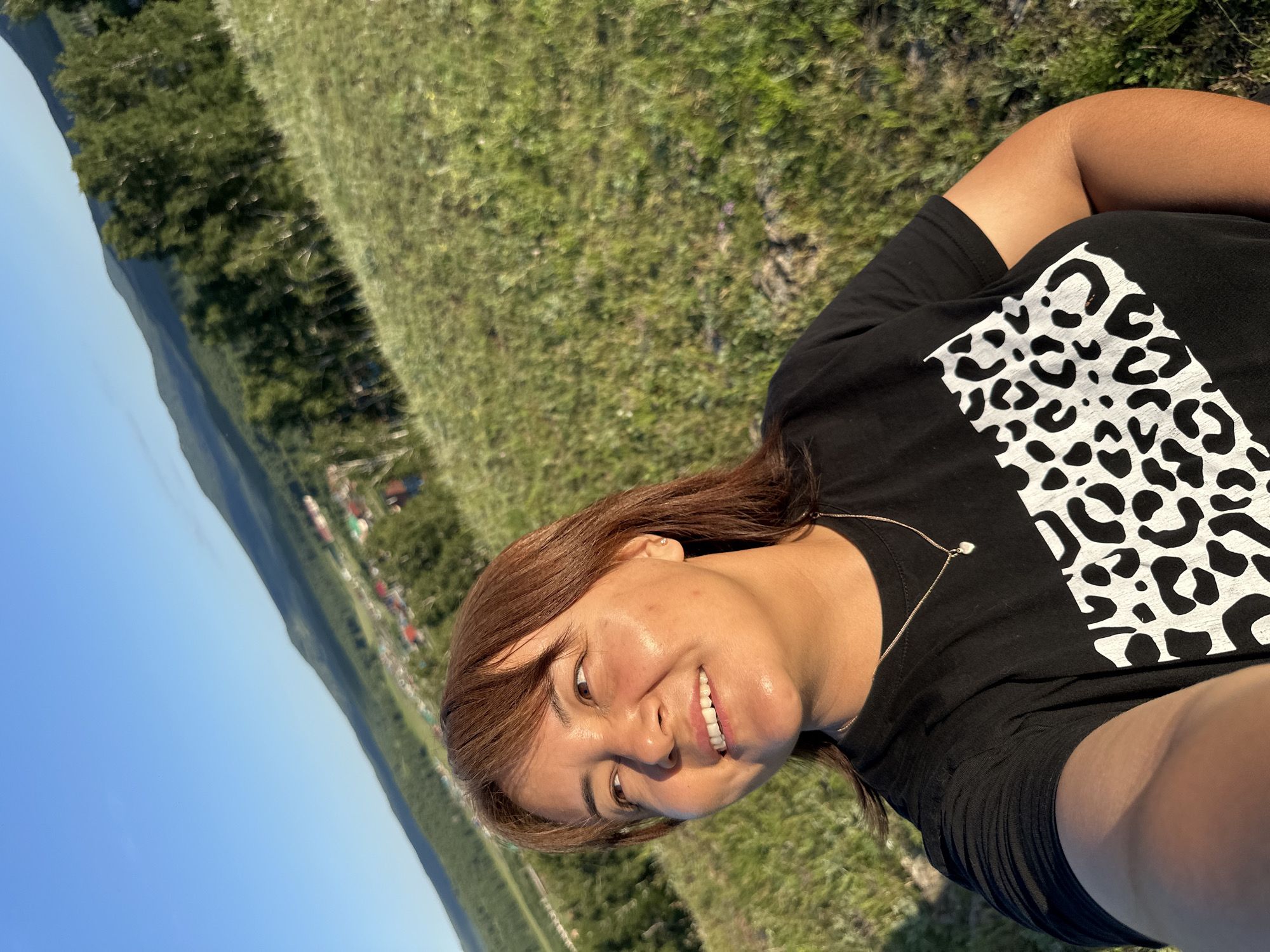 A woman with brown hair and brown eyes is pictured from the chest up, in a selfie taken outdoors. She is wearing a black t-shirt with a white leopard print design across the front and a thin gold necklace with a small heart pendant. She is looking off to the left, with a slight smile. The background consists of a grassy hill sloping downwards, with a village and distant green hills under a clear blue sky. The sun is shining, casting a warm glow on her face.