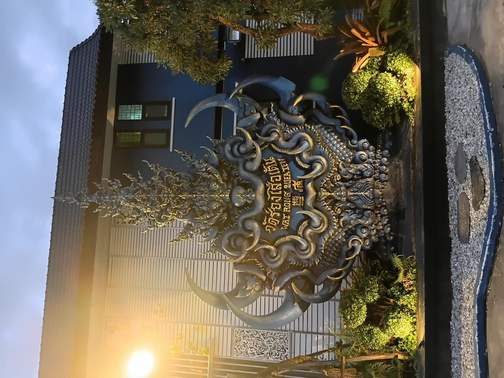 The image shows the entrance to Wat Rong Suea Ten in Chiang Rai City Municipality, Thailand. The focal point is a large, ornate sculpture at the entrance. The sculpture is intricately detailed, featuring what appears to be a dragon or mythical creature in shades of dark gray and gold. It is illuminated from below by spotlights, casting a warm glow on the surrounding greenery. Behind the sculpture, a building with blue walls and a dark tiled roof is visible. The building has several windows, some with patterned glass. The sky is overcast, suggesting it is either early morning or late evening. The ground in front of the sculpture is covered in white gravel, with a few decorative stones and a blue-lined border. To the left and right of the sculpture are small, well-maintained gardens with lush green plants, and a tree with a visible trunk. A bright, warm light source, possibly a streetlamp, is prominent on the left side of the image, illuminating the facade of the building and the trees.