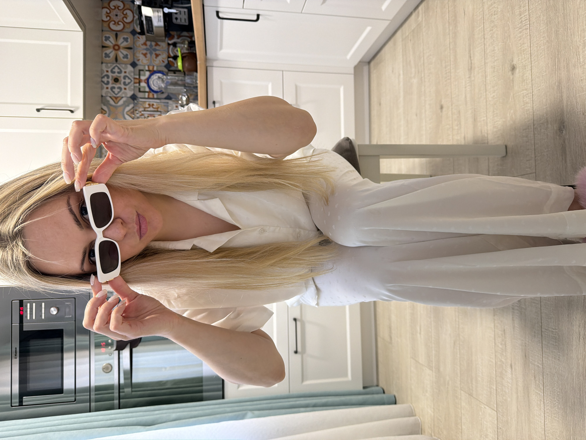 A young woman with long blonde hair, wearing a white button-up shirt and possibly matching trousers, is pictured holding white-framed sunglasses near her face, looking directly at the camera. Her hands, with manicured nails, are positioned as if she is about to put on or take off the eyewear. She has a neutral to slightly expressive look.
The scene is set in what appears to be a modern kitchen in Votkinsk, Russia. To the viewer's left, a stainless steel microwave and oven are visible, with the brand "Hansa" clearly legible on the microwave's panel. A light blue curtain or fabric is seen in the foreground on this side. To the viewer's right and behind the woman, white kitchen cabinets are present. A decorative tiled backsplash, featuring patterns in shades of blue, grey, and brown, adds a splash of color to the background. The floor is made of light-colored wooden planks. Part of a chair with a grey cushion can be seen behind the woman. The lighting suggests it is daytime, and the overall atmosphere is casual and relaxed within a home setting.