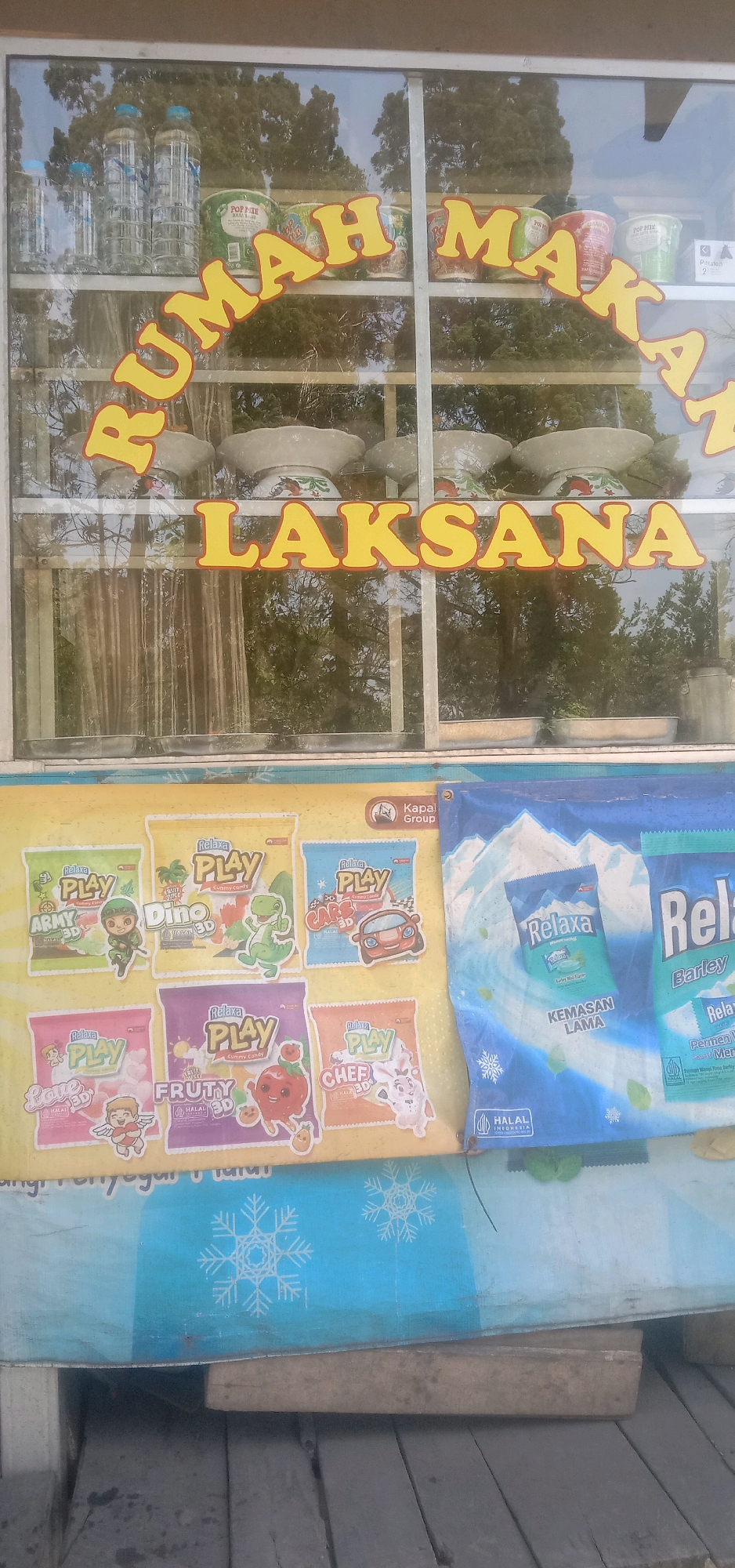 The image captures a storefront display, likely in Palasari, Indonesia, featuring a variety of products. At the top, visible through a glass window, are shelves stocked with bottled water, cans of instant noodles labeled "POP MIE," and other packaged goods. A large yellow sign arching across the window reads "RUMAH MAKAN" (Restaurant) and "LAKSANA" in bold letters. Below this, several white ceramic bowls are displayed.
The lower portion of the image showcases two large banners. The left banner is yellow and features an assortment of colorful packets of "Relaxa PLAY" gummy candies, each depicting different characters like soldiers, dinosaurs, cars, cherubs, fruits, and cartoon animals. The right banner is blue and features images of snowy mountains and packages of "Relaxa" mints and "Relaxa Barley" products. The bottom of the image shows wooden planks, suggesting the display is at ground level or on a raised platform. A blue tarp with snowflake motifs is draped below the banners. The overall scene suggests a small, local eatery or convenience store.