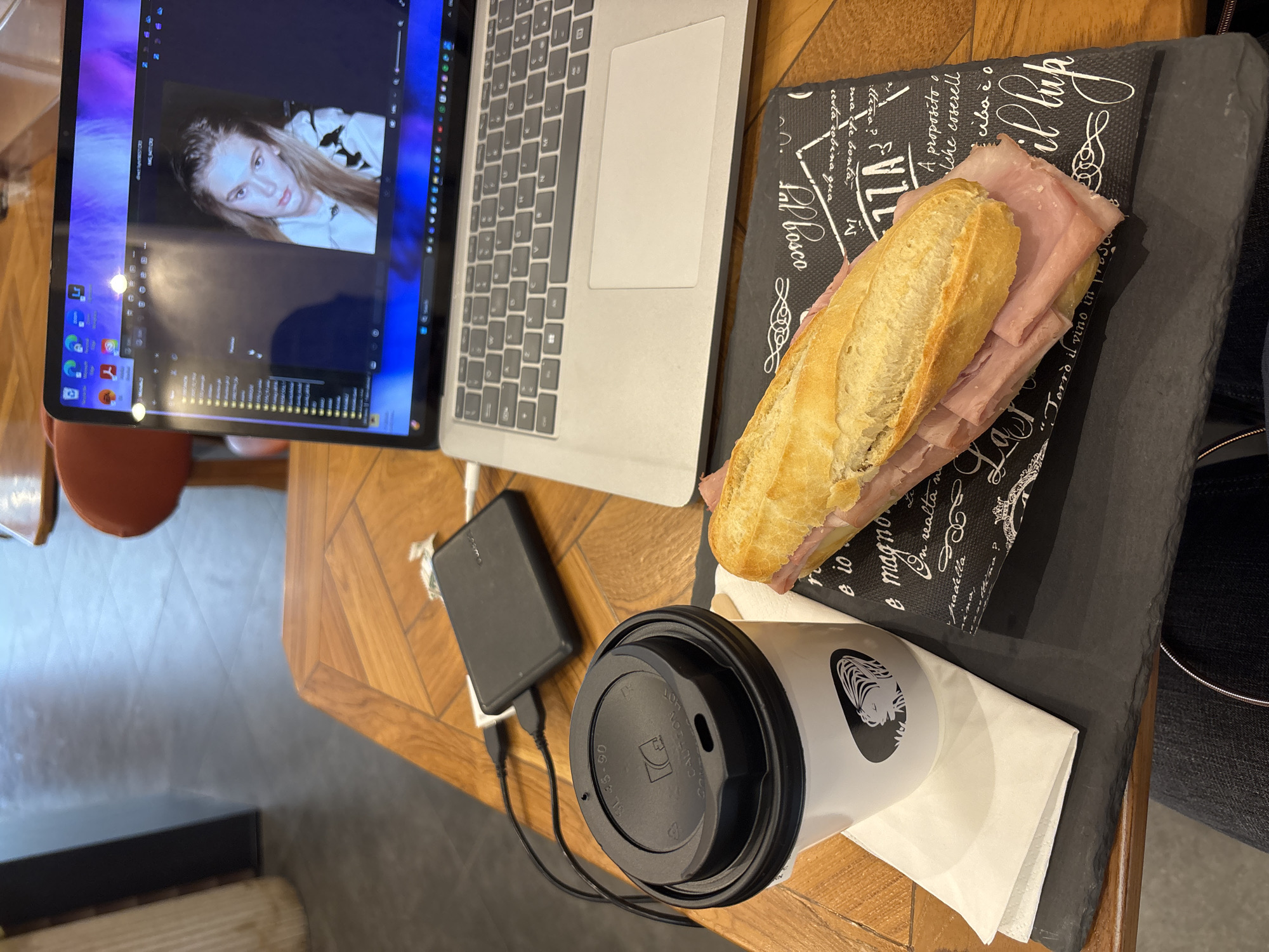 The image presents an overhead view of a work or leisure setup on a wooden table with a geometric pattern, likely within an indoor environment such such as a cafe or home in Milan, Italy.
A silver laptop, possibly a MacBook, is open on the left. Its screen prominently displays a woman's face with long, light brown hair, suggesting a video call or image editing session. Visible on the screen are icons for applications like Zoom, Lightroom, and Microsoft Word, along with what appears to be an audio equalizer or waveform at the bottom.
To the right of the laptop, a sandwich made from a sliced baguette filled with ham or cold cuts is placed on a black rectangular placemat. The placemat is adorned with white decorative script and phrases in Italian, including "il lupo," "A proposito o," "Padella," "magno," "in realta," and "Pal Bosco."
Below the sandwich is a white takeaway coffee cup with a black lid. The lid displays the warning "CAUTION HOT" and "SL 45-90," while the cup itself features a distinct black logo, possibly a stylized mermaid or siren. Adjacent to the coffee cup, a black power bank is connected by USB cables, indicating a device is being charged. A white paper napkin rests underneath the cup.
The scene conveys an individual engaged in work or personal activity, accompanied by a meal and a drink.