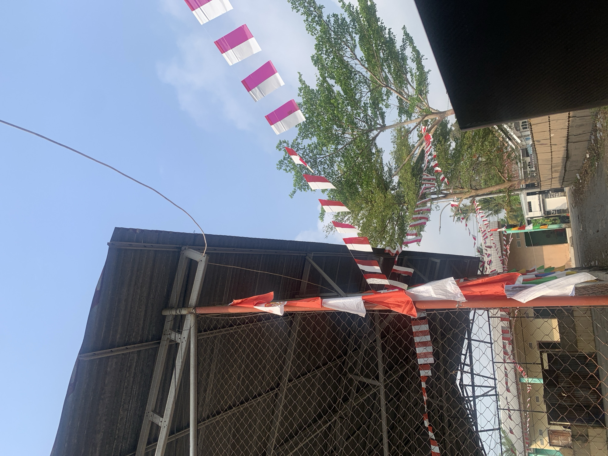 The image captures an outdoor scene with elements suggesting a celebration or patriotic event, likely in Palembang, Indonesia, given the red and white flag decorations. In the foreground, a metal-fenced structure with a corrugated metal roof is visible. A reddish-orange pole stands vertically, adorned with red and white flags, some of which are draped or fluttering. A string of red and white pennant flags stretches across the scene, some appearing to be Indonesian flags. In the background, a tree with lush green foliage is prominent, with more red and white flags strung between poles and branches. Beyond the tree, buildings with tiled roofs are visible, hinting at a residential or community area. The sky is clear and blue, suggesting daytime. The overall impression is one of festivity and decoration, with a clear emphasis on the colors red and white.