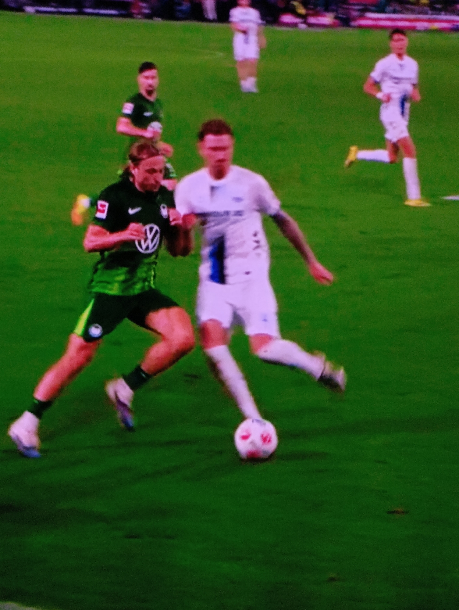 A soccer game is in progress on a green grass field. A player in a green and black uniform with the number 9 on the left sleeve, marked with the Volkswagen logo, is actively involved in the game. He is positioned close to the ground and appears to be challenging for the ball. A second player, dressed in a white uniform with a blue stripe on the side, is actively kicking the soccer ball. The ball is located between the two players. A third soccer player wearing a green and black uniform can be seen in the background slightly behind the first player. In the upper right corner, another soccer player wearing a white uniform is running. There are additional indistinct figures on the field in the background. The city of Plovdiv, Bulgaria, is not identifiable in the image.