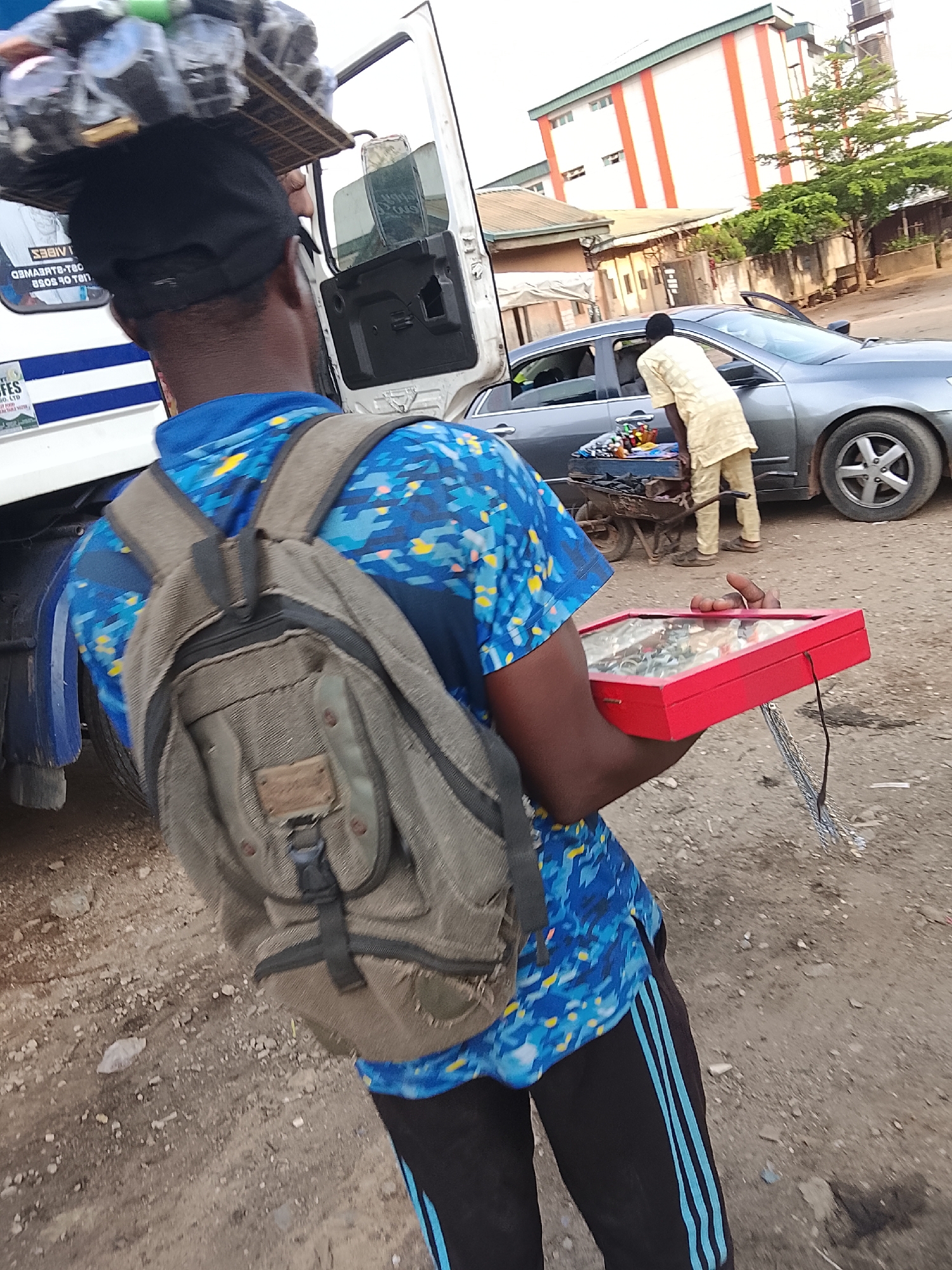 A street vendor stands on a dirt road in Abuja, Nigeria. He balances a large tray of items on his head, wears a backpack, and holds a red jewelry box displaying silver-colored necklaces. He faces toward a parked gray car and a man examining wares on a cart behind it. A large white truck stands to the vendor’s left, with buildings and trees in the background.