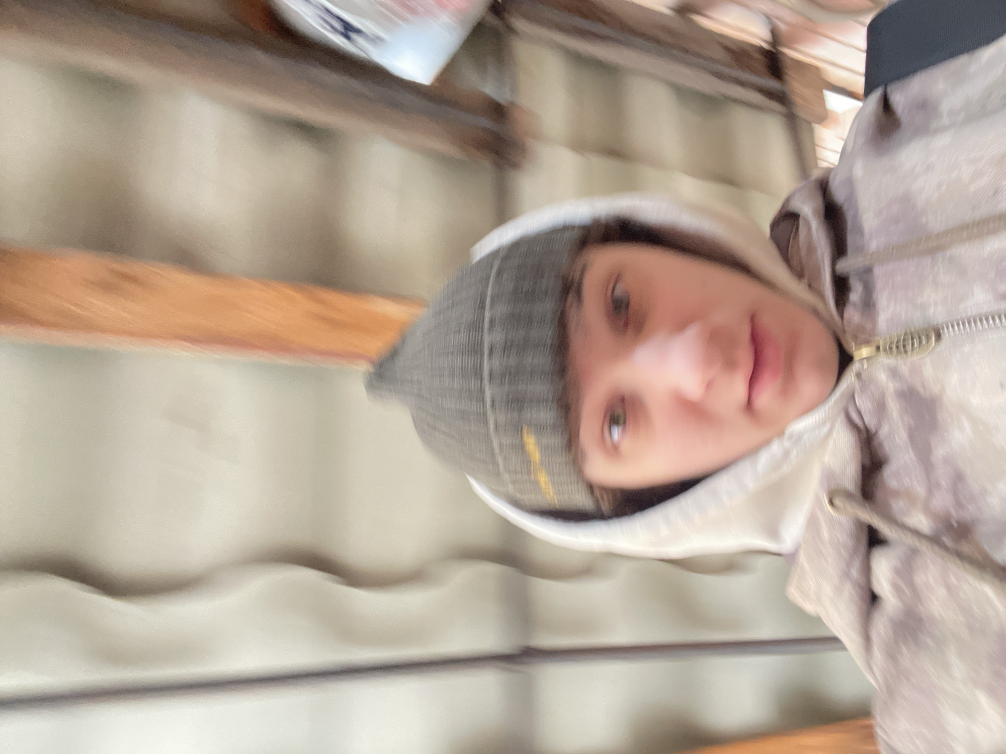 The image displays a close-up, upward-angled portrait of an individual, possibly a selfie, taken in what appears to be an indoor or semi-enclosed setting. The photo is rotated 90 degrees clockwise.
The main subject is a young person, whose face is partially visible. They wear a grey knitted beanie with a thin yellow stripe or design, a light-colored hooded sweatshirt, and an outer jacket with a subtle camouflage or patterned texture in light brown and grey tones. A zipper is visible on the jacket. Their right eye (from the viewer's perspective), part of their nose, and lips are discernible, with their gaze directed towards the camera, conveying a neutral to relaxed expression. Their dark hair is visible beneath the beanie.
The background suggests an older or utilitarian interior space. Two prominent wooden beams or planks, light brown in color, span horizontally across the frame. Behind these, a light grey or off-white corrugated surface, possibly a ceiling or wall, is visible. In the upper left corner, a blurry white object with a hint of blue, possibly a package or container, rests on one of the upper beams.
The setting, with its visible wooden beams and corrugated material, could be indicative of a garage, workshop, attic, or a similar functional building. While the provided context states Krasnoyarsk, Russia, there are no specific visual cues within the image itself to confirm this location. The image quality has some blur, particularly in the background, which could be due to movement or focus. No explicit text is clearly legible in the photograph.