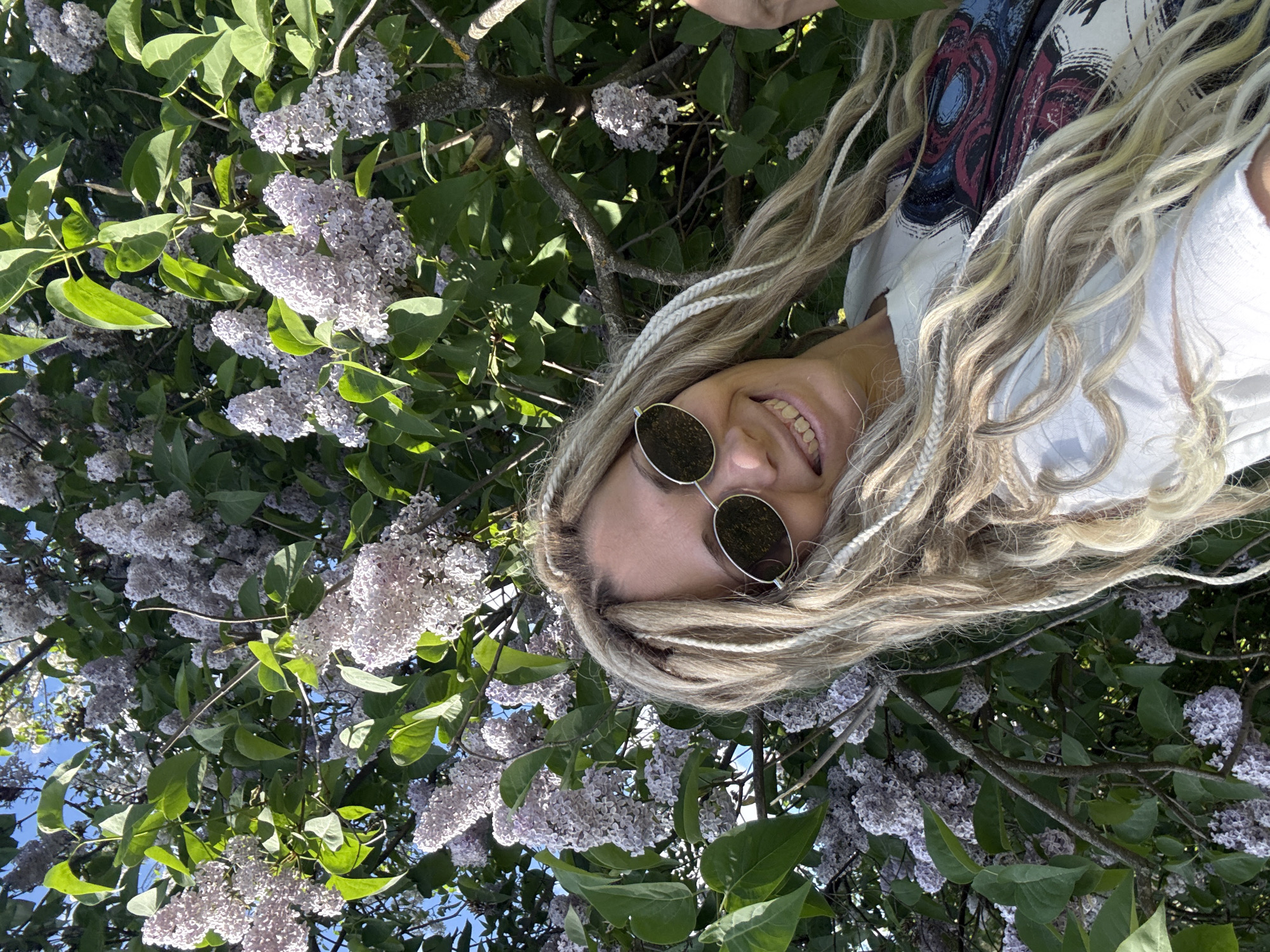 A person with light-colored dreadlocks and round sunglasses smiles amidst blooming lilac bushes. The foliage and flowers dominate the frame, with glimpses of blue sky visible between the branches. The location is the city of Шебунино, Russia. The subject is wearing a white top.