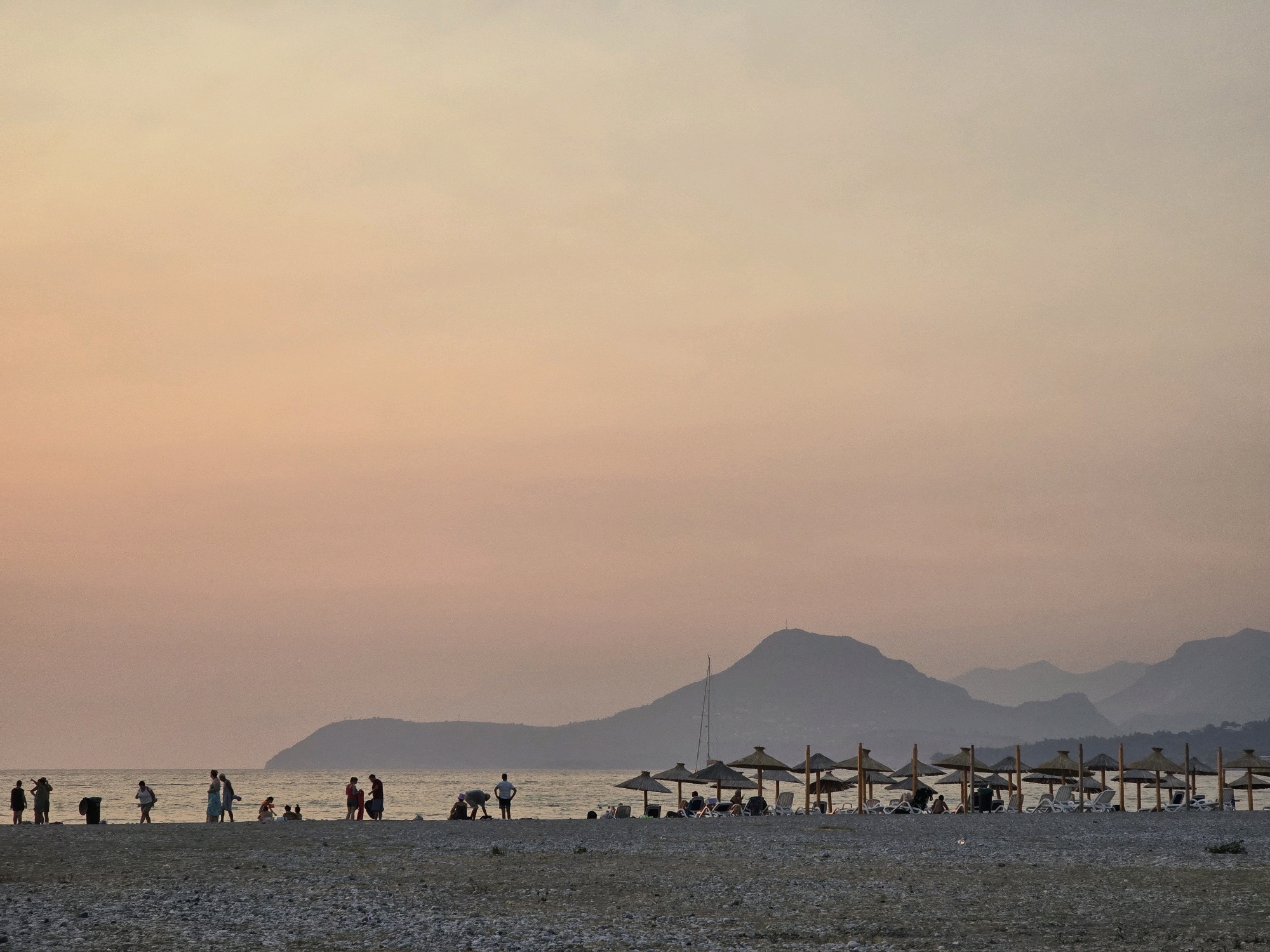 This is a wide shot of a pebbly beach in Bar, Montenegro, at sunset. The sky is a gradient of soft orange and pink, transitioning to a muted gray towards the horizon. In the distance, silhouetted mountains form a layered backdrop.
On the beach, a line of thatched umbrellas with lounge chairs is visible, suggesting a resort or public beach area. A few people are scattered along the shoreline, some walking and others sitting. Further left, a group of people are gathered near the water's edge, their forms indistinct against the soft light. The sea is calm, reflecting the muted colors of the sky. The overall impression is one of tranquility and an end to a day at the beach.
