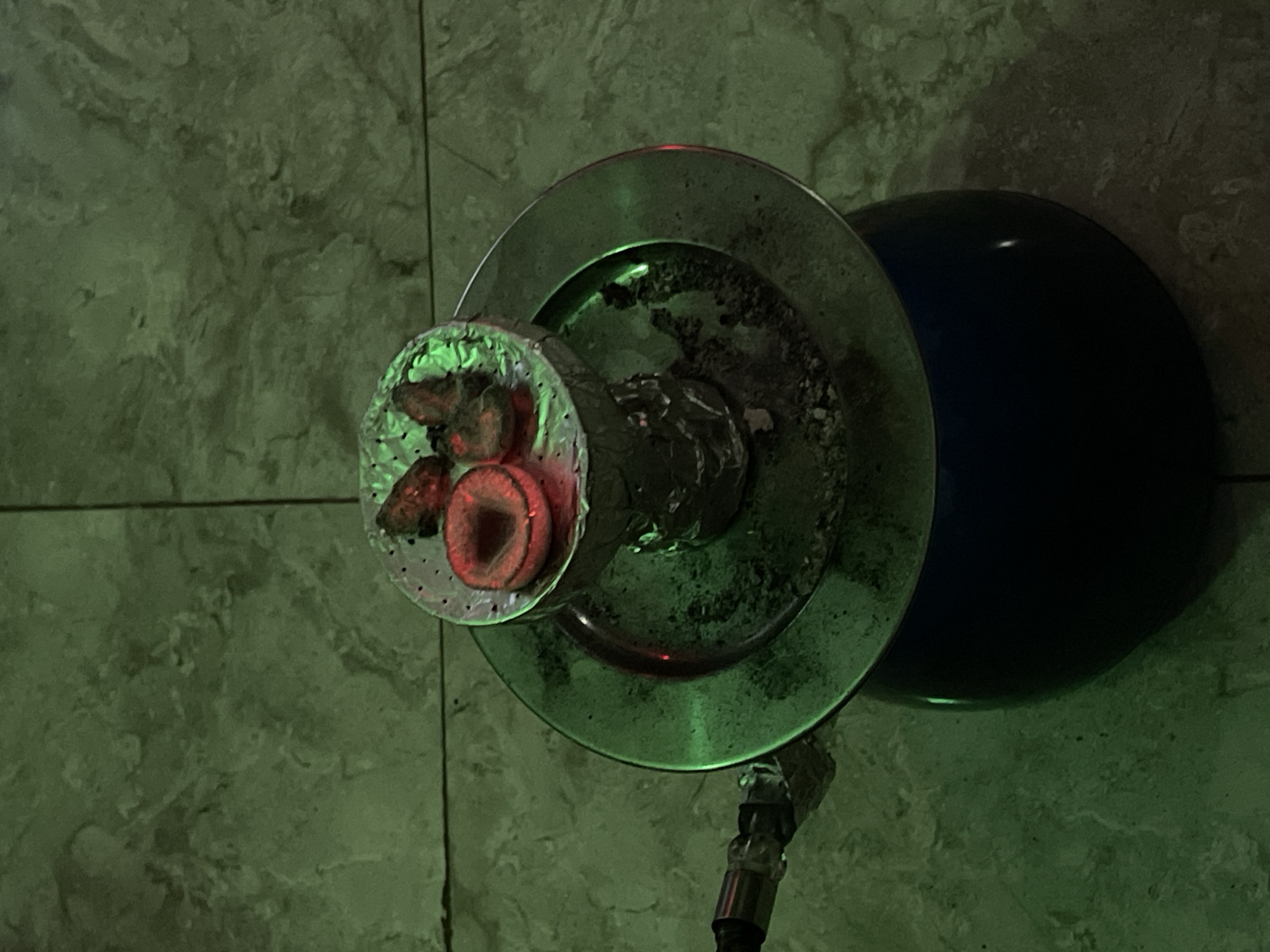 A top-down view shows a lit hookah on what appears to be a tiled surface. The hookah bowl is filled with hot coals, glowing red. The hookah base is dark blue, and a metal tray sits beneath the bowl, containing burnt tobacco. A hose is connected to the base. The scene is lit with a green-tinted light. The location, Kuchingoro, Nigeria, is not ascertainable from this image.