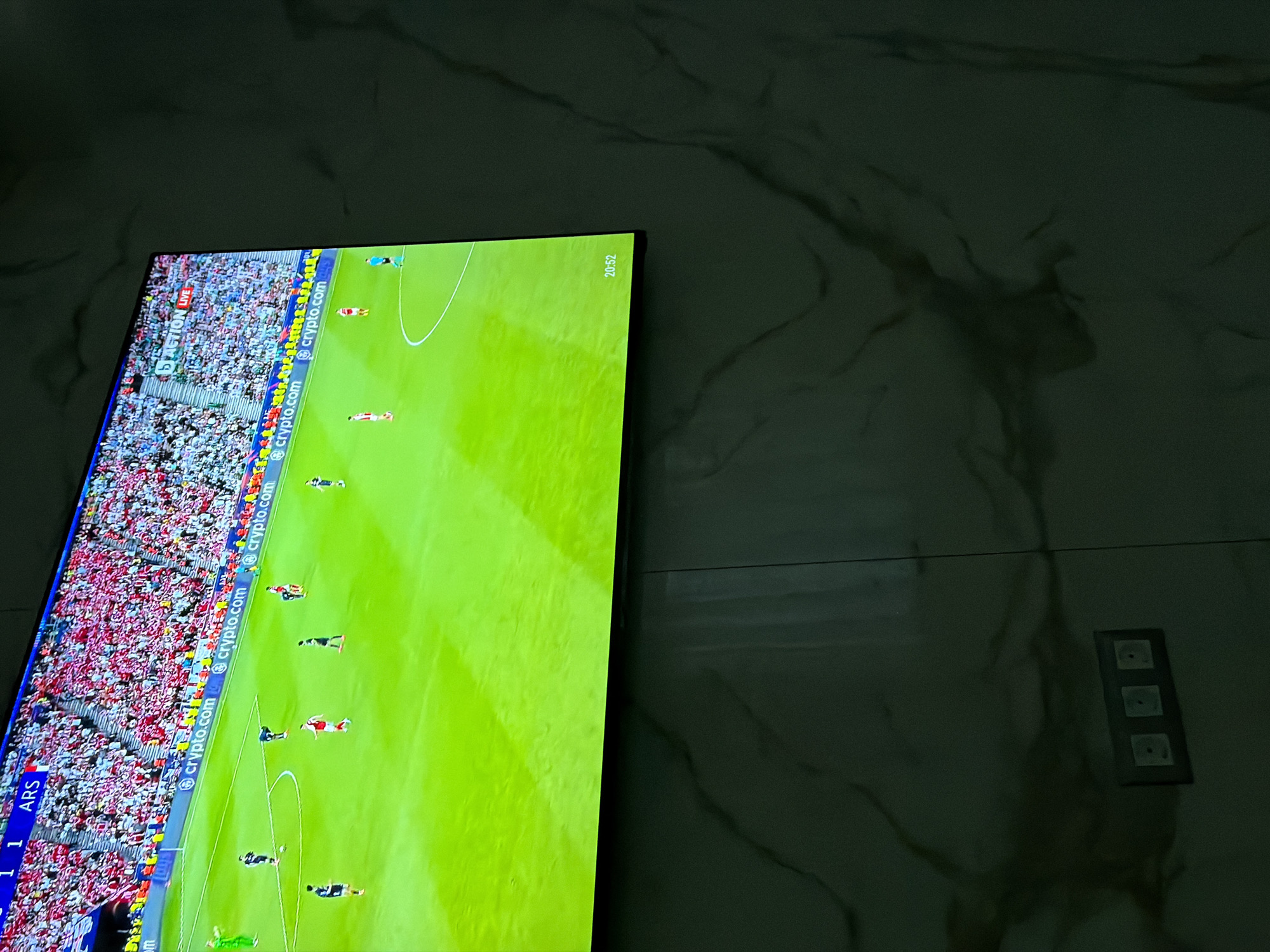 The image shows a television screen displaying a soccer game. The game appears to be between Arsenal (ARS) and another team, with the score tied at 1-1. The match seems to be taking place in a stadium with a large crowd. The television is mounted on a wall that has a marble-like finish. There are three light switches on the right side of the screen. The time shown on the screen is 20:52. There is no information in the image to indicate the location is in Asenovgrad, Bulgaria.