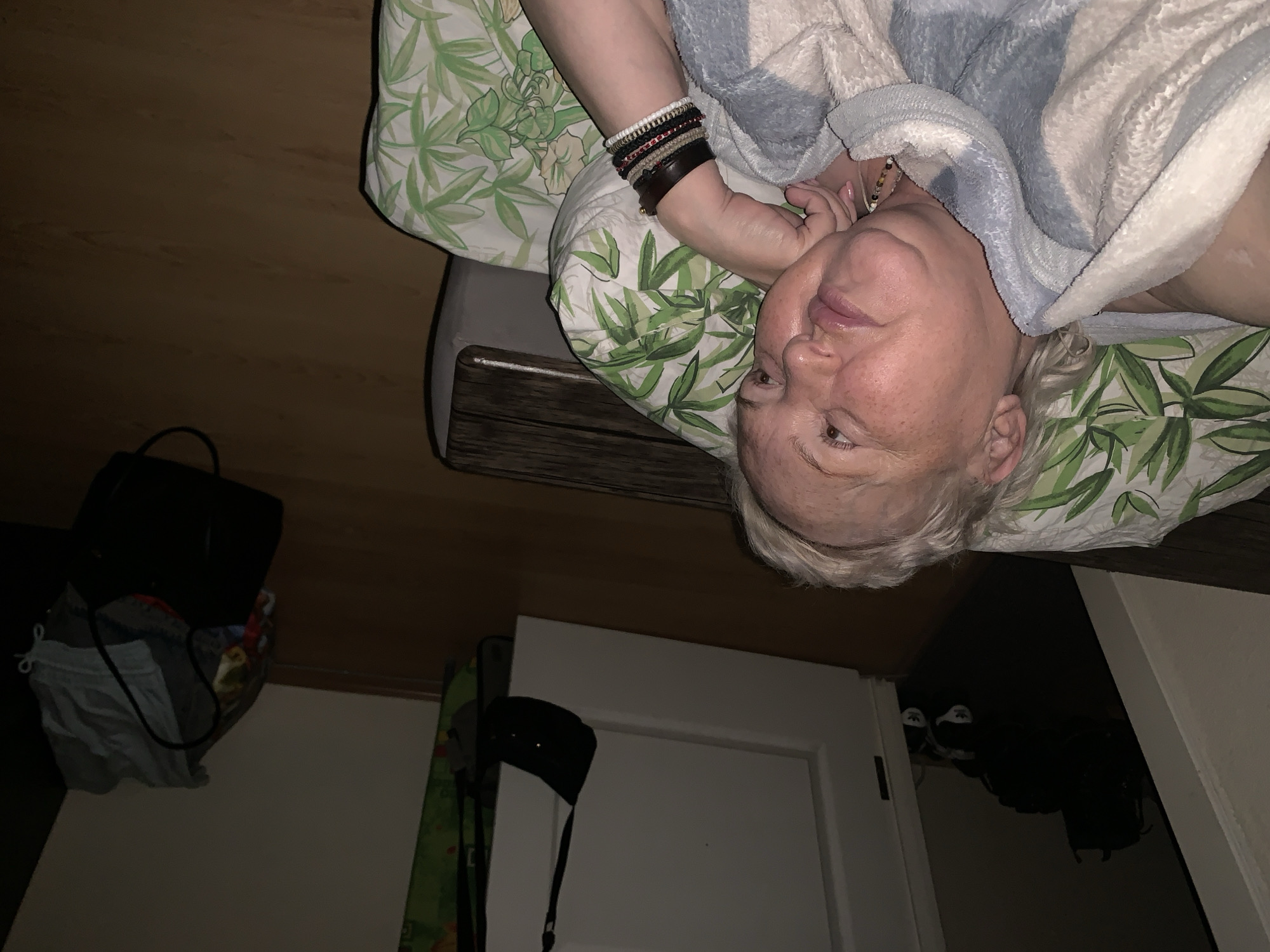 An elderly woman is reclining on a patterned pillow, covered with a soft, blue and white blanket. She is wearing several bracelets on her left wrist. The background shows an interior scene with a partially visible door, a black bag, and other items stored in a dark space. The location of this image is not specified.