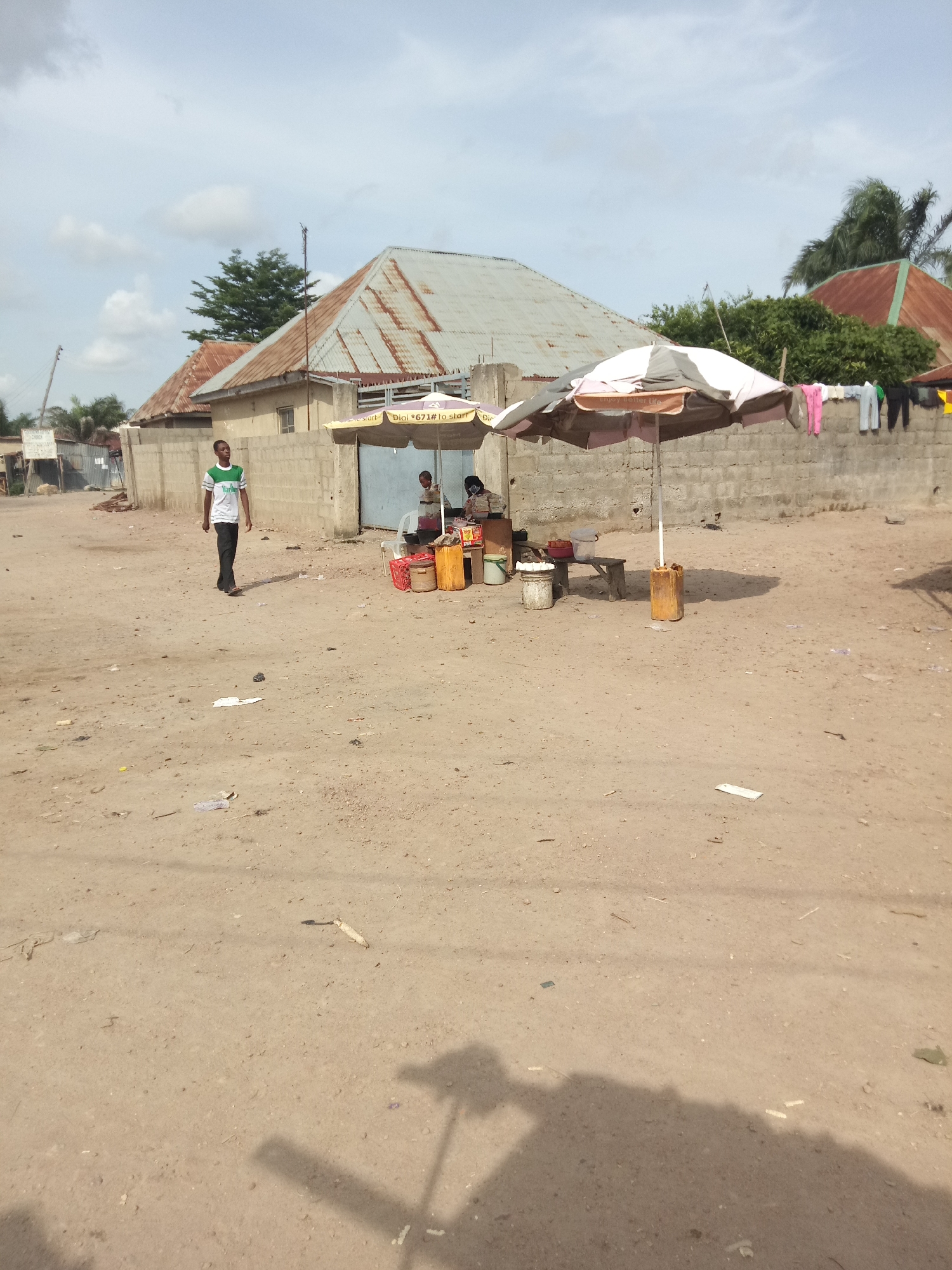 A man in a white and green striped t-shirt and black pants walks across a dusty, unpaved street. In the background, two people are standing near a market stall under a large umbrella. The stall has a table with some items on it and a few buckets. A wall made of concrete blocks runs behind the stall, and laundry is hanging on it. Several corrugated metal-roofed buildings are visible further in the background. The sky is partly cloudy.