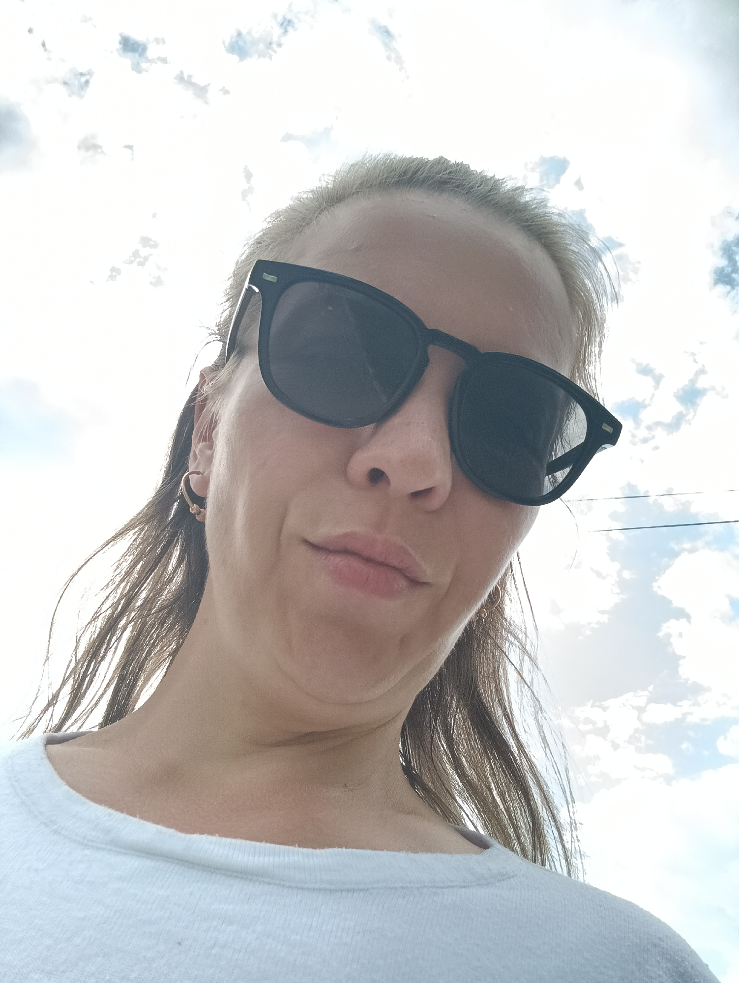 A close-up, low-angle selfie of a person wearing sunglasses and a white t-shirt is captured against a bright, cloudy sky. The person's face is tilted upwards, and their expression is neutral, with their lips slightly parted. The background consists of a sky filled with white, fluffy clouds and hints of blue. The image has a slight sepia tone, and there are visible strands of the person's hair blowing in the wind. The selfie appears to have been taken outdoors, possibly on a sunny day.