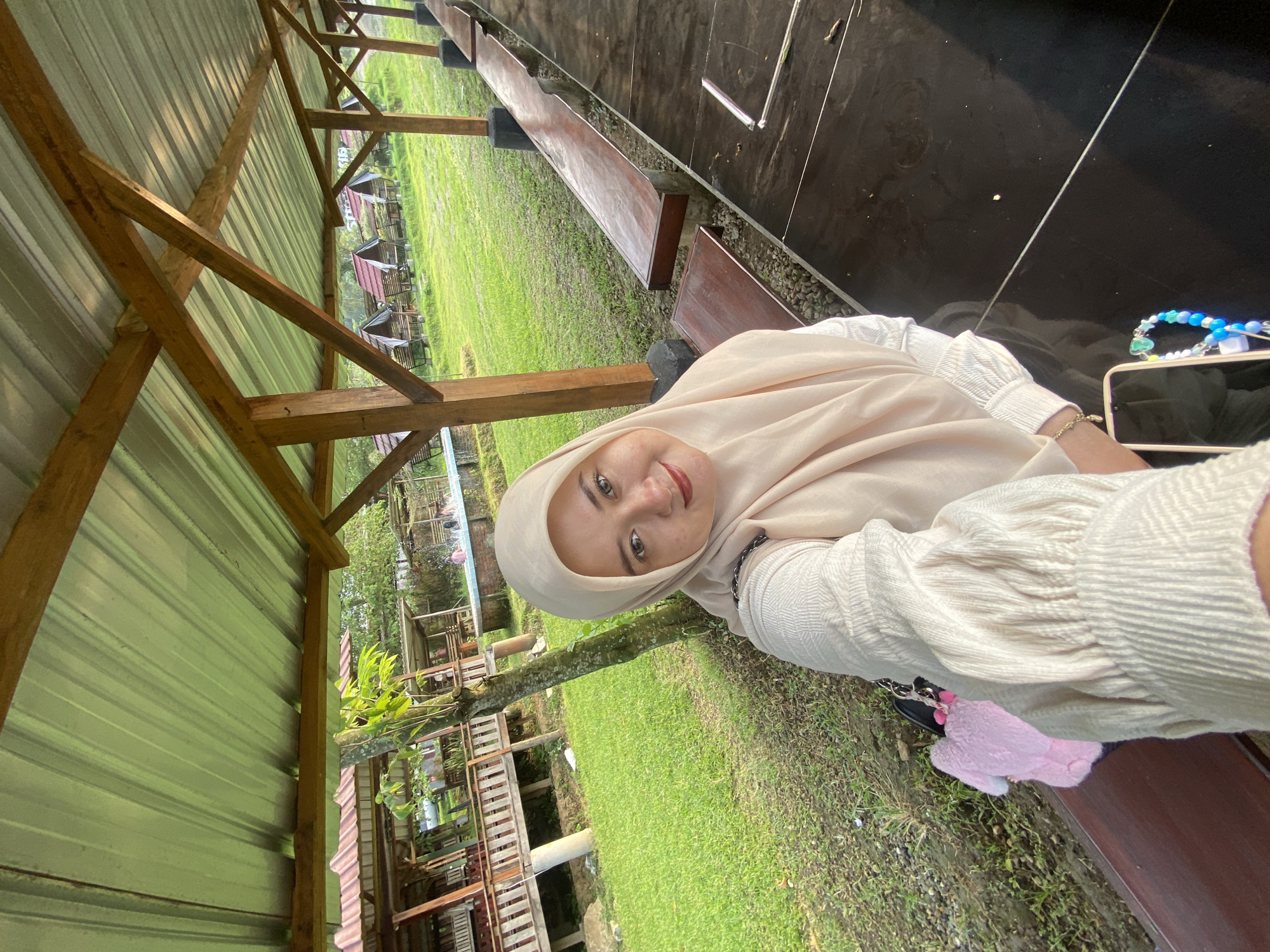 A woman wearing a light-colored hijab and blouse is seated on a bench, seemingly taking a selfie. The location appears to be an outdoor recreational area in Penanggungan, Indonesia, characterized by wooden structures, green grass, and a metal roof overhead. A mobile phone and a beaded bracelet are visible next to her. There are other benches alongside.