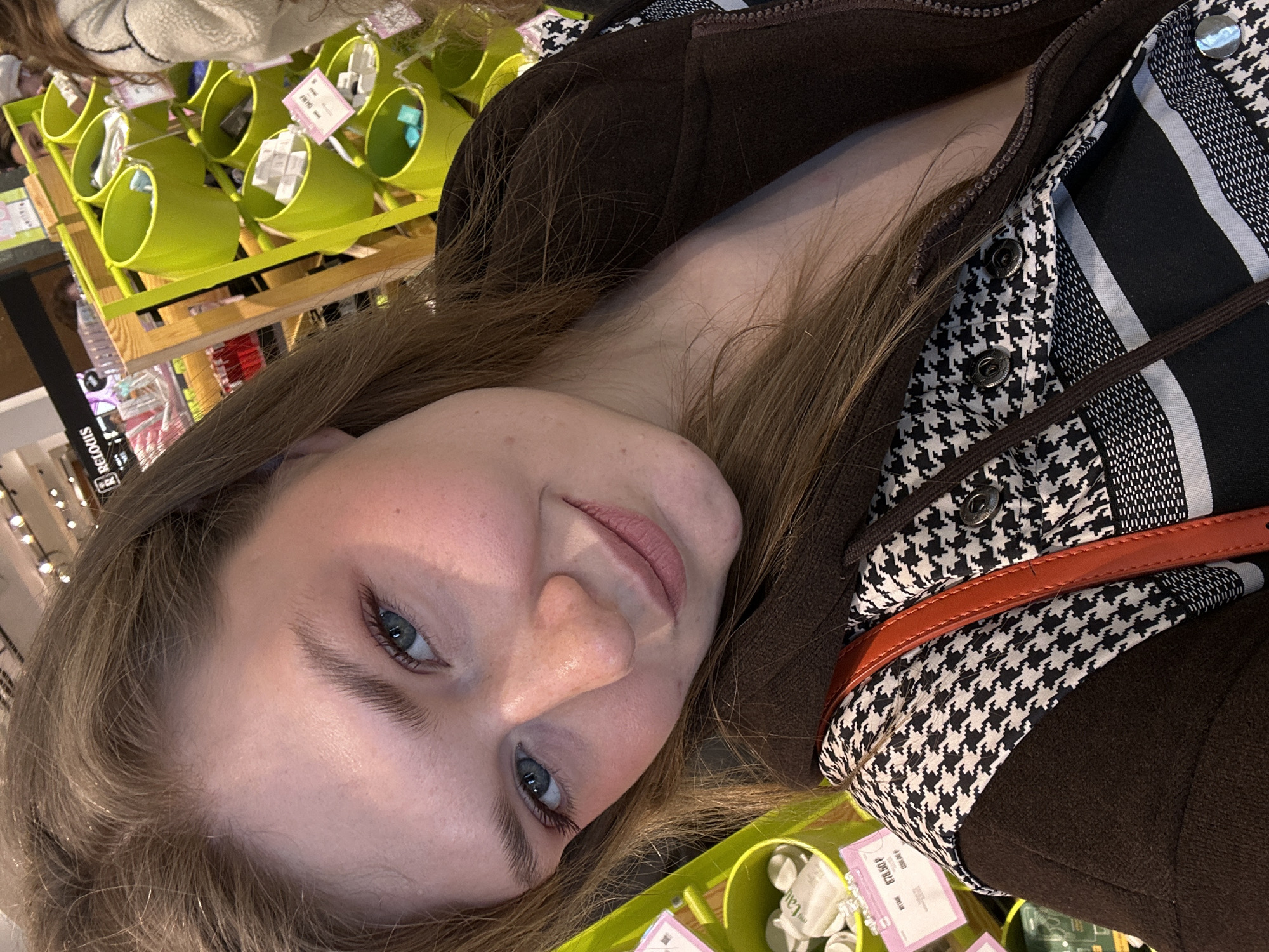 The image presents a close-up selfie of a fair-skinned female, appearing young to middle-aged, with light brown hair and blue eyes, looking directly into the camera. Her mouth is slightly upturned at the corners. She is wearing a dark brown zippered jacket over a black and white patterned shirt, which features a houndstooth design on the upper portion and horizontal stripes on the lower section. An orange strap, consistent with a purse or bag, is visible across her left shoulder.
The background depicts an indoor retail environment, specifically a cosmetics or beauty store in Kazan, Russia, indicated by a visible "RELOIS" sign. The store's interior includes light wooden shelving units holding multiple lime green cylindrical buckets. These buckets contain various small, packaged products, predominantly white and light blue. Pink price tags, displaying Cyrillic text and prices, with one visible showing "878.50 Р", are attached to the shelving. Blurred, partial figures of at least two other individuals are discernible in the background. The scene is illuminated by overhead artificial lighting.