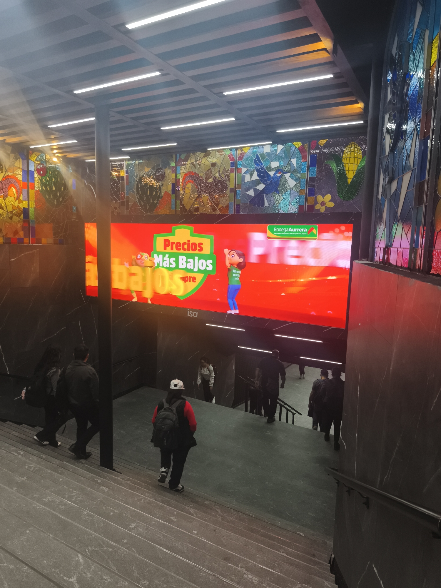 This indoor photograph captures a multi-level public space, likely a metro station or a large building entrance in Mexico City, Mexico. The scene is characterized by a blend of modern architecture and vibrant artistic elements.
Dominating the upper center is a large, brightly lit LED screen displaying an advertisement for "Bodega Aurrera," a Mexican supermarket chain. The screen features a cartoon character and bold text in Spanish: "Precios Más Bajos siempre" (Lowest Prices always), with the Bodega Aurrera logo and slogan "La supercampeona de los precios bajos" (The super champion of low prices) in the upper right. Below the screen, the text "isa" is visible on a dark panel.
Above and flanking the LED screen are expansive, colorful stained-glass murals that adorn the upper walls. These artworks depict a variety of designs, including a blue bird, a yellow ear of corn, geometric patterns, and abstract forms in vivid yellows, blues, greens, and reds.
The architectural elements include dark, polished stone walls, possibly marble or granite, with subtle white veining, and a sturdy dark pillar supporting the structure. Wide, grey stone staircases descend through the space, accompanied by dark handrails. The ceiling features a grid pattern embedded with linear fluorescent lights, providing bright illumination.
Several individuals are present, actively descending the stairs. In the foreground, a person wearing a red top, a white cap, and a black backpack is seen moving downwards. Other figures are visible further along the stairs and on a lower landing, all appearing to be navigating the space. The setting suggests a public transportation hub or a busy commercial building.