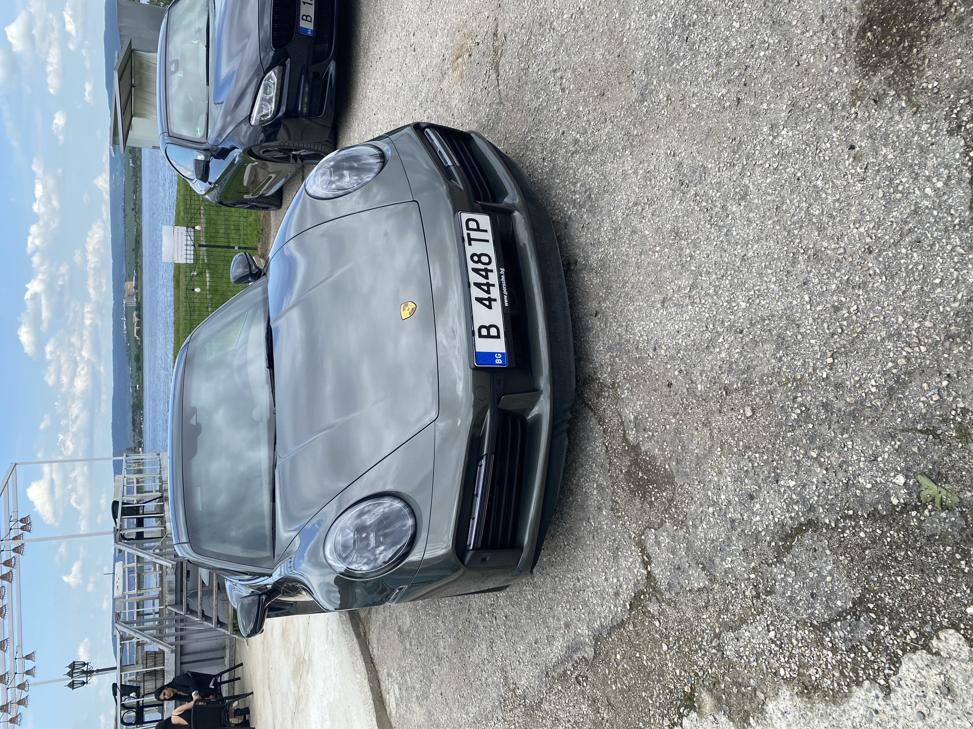 The image shows two parked cars. The car in the foreground is a gray Porsche with the license plate "B 4448 TP," indicating registration in Bulgaria. The second car behind the Porsche is black, but its license plate is only partially visible, reading "B 1." The vehicles are parked on a concrete or asphalt surface. In the background, there is a view of water, possibly a lake or river, and a hillside. To the left, there is an outdoor structure with hanging lamps, and two people are seated at a table. The location is Konstantinovo, Bulgaria.