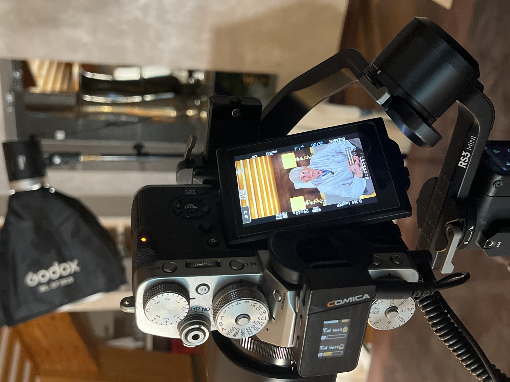 The image depicts a close-up view of a camera setup, likely for recording a video. A silver Fujifilm camera with various dials and buttons is mounted on a black gimbal stabilizer, identified by the "RS3 MINI" text. The camera's screen displays a recording of an elderly man with grey hair, wearing a light blue lab coat or smock, and holding a pen. He appears to be in a studio setting, with a softbox light from a "Godox" brand visible in the background. The overall lighting suggests an indoor environment, possibly a studio or a well-lit room. There are no discernible elements in the image that would indicate the location as Kazan, Russia.