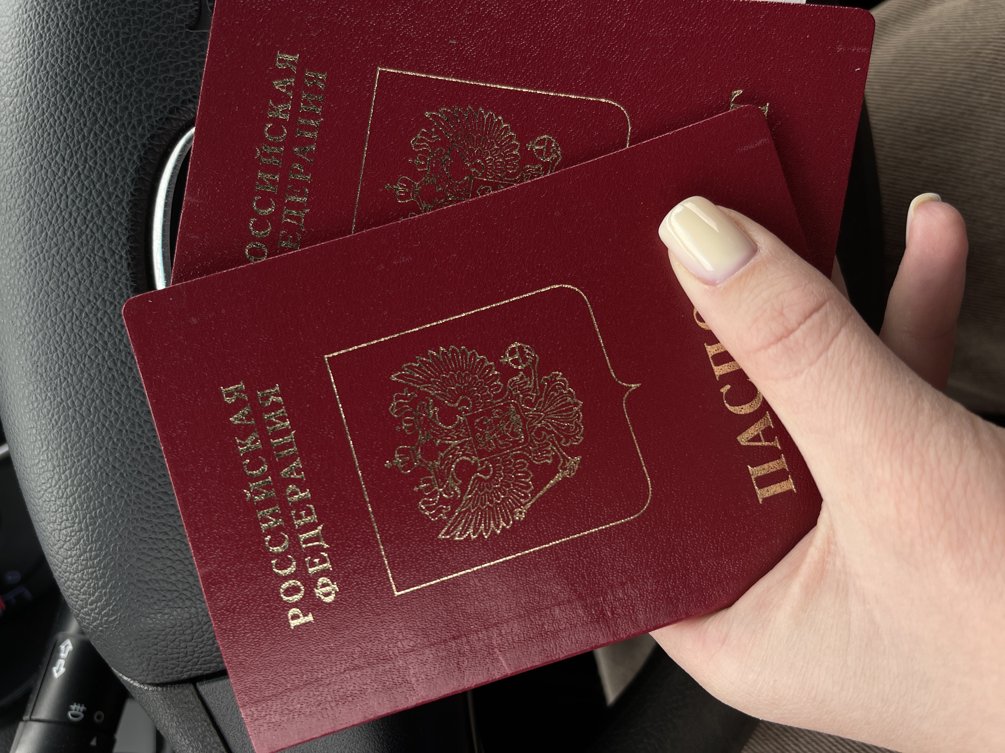 A close-up shot shows a fair-skinned hand with manicured nails holding two maroon passports, identified by the gold text as Russian Federation passports. The passports bear the emblem of Russia in gold foil. The hand is positioned over a dark-colored steering wheel. The exact location is not specified and cannot be derived from the image provided.
