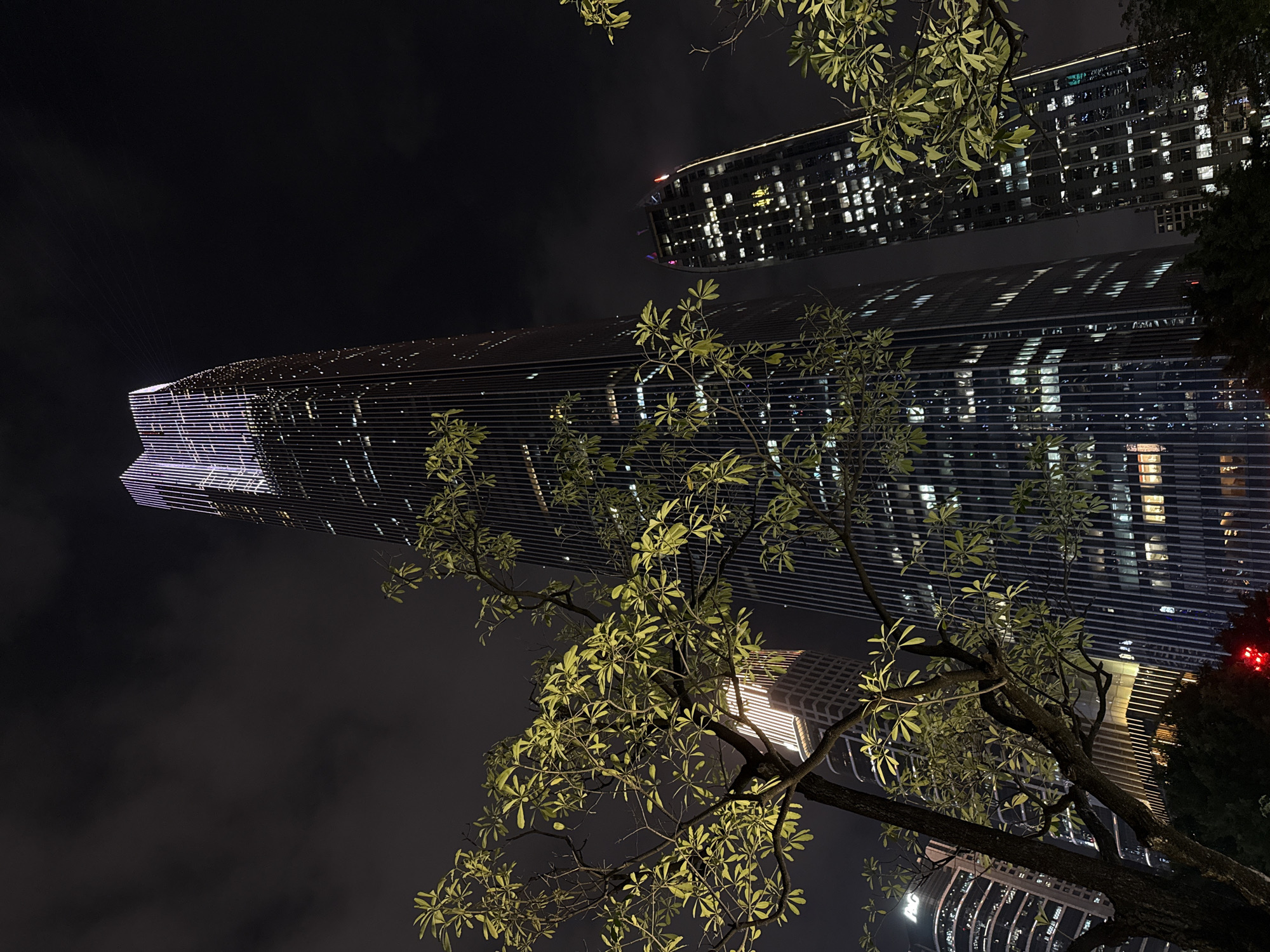 The image shows two skyscrapers at night, located in Tianhe District, China. A tree with illuminated green leaves is in the foreground, partially obscuring the view. The buildings are lit with numerous windows, suggesting offices or residential spaces. The sky is dark, and visibility is low due to the darkness and atmospheric conditions.