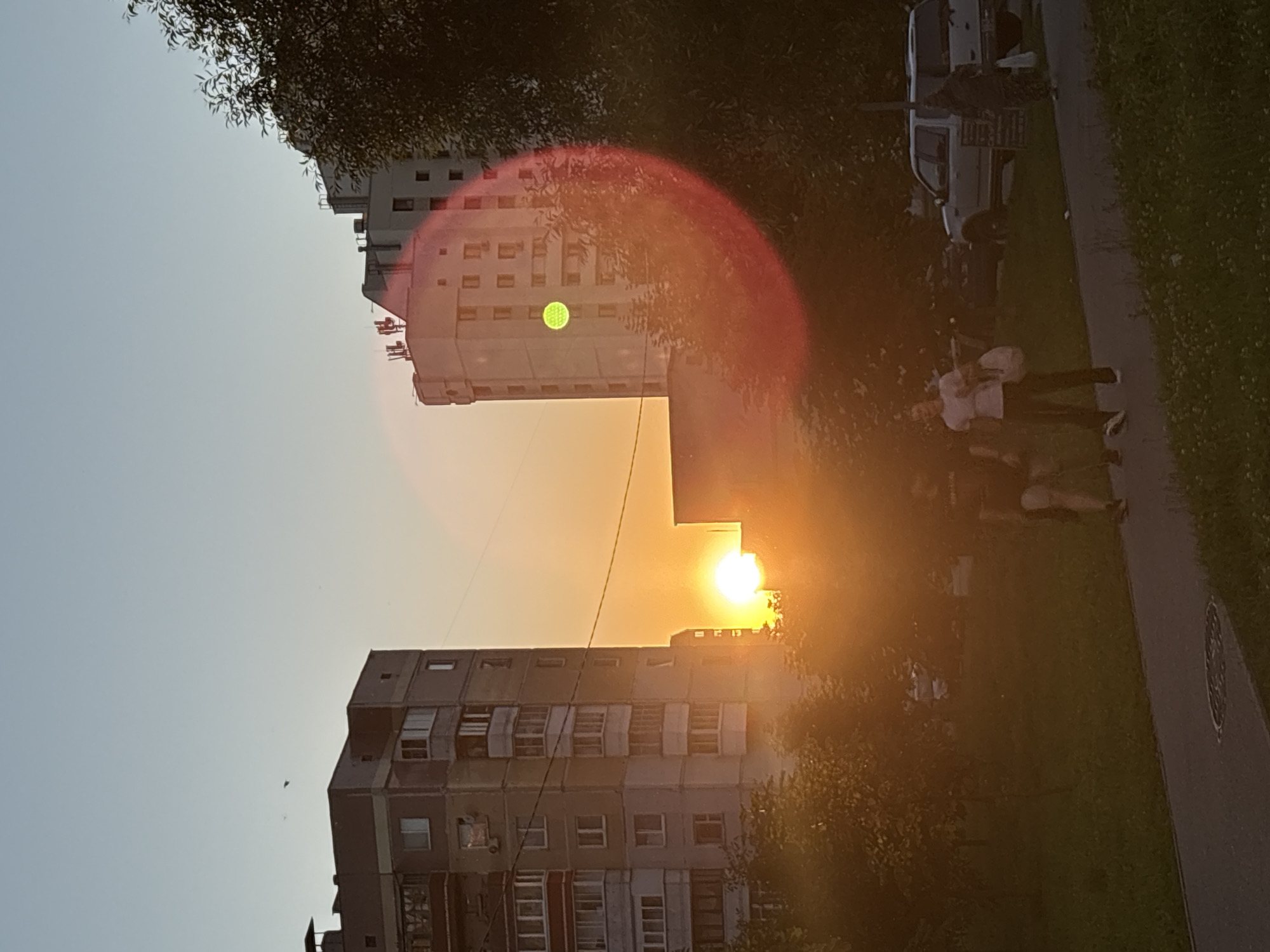 This media file depicts an outdoor scene at dusk in Vitsebsk, Belarus. The primary subjects are two apartment buildings, one tall and light-colored, the other shorter and darker. A bright sun is setting, casting a warm orange glow across the sky and reflecting off the buildings. The sun is partially obscured by the roofline of the taller building, creating a lens flare effect with a red and orange halo.
In the foreground, to the right of the darker building, is a sidewalk and a grassy area. A few people are visible walking on the sidewalk, appearing as silhouettes against the light. One person in the foreground is wearing a white t-shirt and dark pants. There are also some vehicles in the background near the sidewalk, one of which appears to be a white van. Trees with dense foliage are present, framing the scene and adding to the sense of an urban park or residential area.
The time of day is clearly dusk, indicated by the low angle of the sun and the warm, atmospheric lighting. The weather appears to be clear, with the sun prominently visible. The presence of apartment buildings and a sidewalk suggests an urban or suburban setting. The overall mood is peaceful and serene, characteristic of a sunset. No visible text is present in the image.