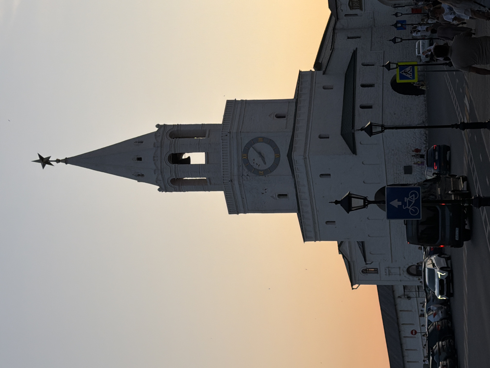 The image shows the Suyumbike Tower in Kazan, Russia, during sunset. The tower, a prominent landmark of the Kazan Kremlin, is a white, multi-tiered structure topped with a red star. A clock face with Roman numerals is visible on one side of the tower. In the foreground, a street is visible with several cars, a van, and pedestrians. Road signs, including one indicating a bicycle path, are also present. The sky is a gradient of pale orange and yellow, indicating the setting sun.