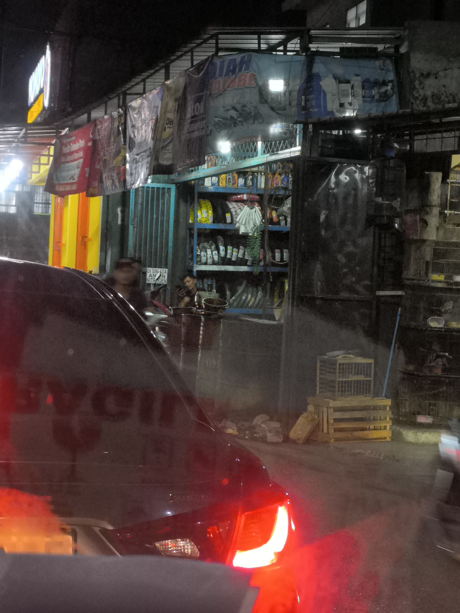 This image depicts a street scene at night in Tangerang, Indonesia, likely near a roadside shop or service area. In the foreground, the rear of a dark-colored vehicle is visible, with its red taillights illuminated, suggesting it is stopped or moving slowly. The shop in the background appears to sell automotive fluids, such as oil, displayed on shelves behind a counter. Several banners hang from the awning of the shop, some advertising products. Two individuals are partially visible within the shop's interior. One person, seen from the chest up, is wearing dark clothing and a hat, and is looking towards the right. Another person is seated further back, also looking towards the right. The environment appears to be an urban area with mixed-use buildings.