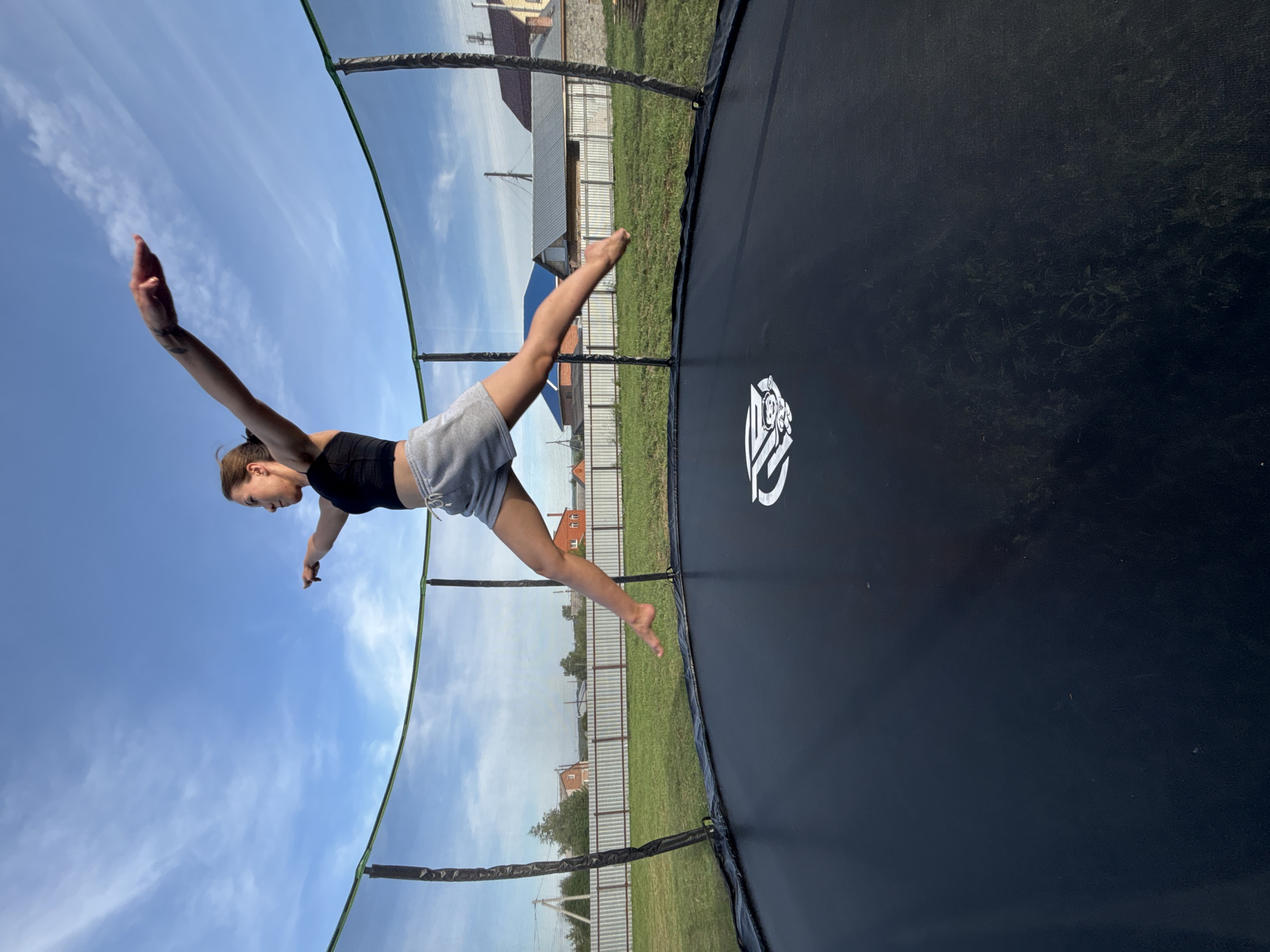 A young woman with brown hair tied in a ponytail is captured mid-air on a trampoline, her body forming a wide, dynamic pose. She is wearing a black crop top and grey shorts. Her arms are extended outwards, and her legs are spread apart in a graceful leap. The trampoline is black with a white logo in the center and is enclosed by a green safety net. The background reveals a rural setting, likely Vishnevka, Russia, with a green lawn, a white fence, and several houses with pitched roofs under a clear blue sky with scattered white clouds. The sun appears to be setting, casting a warm light on the scene.