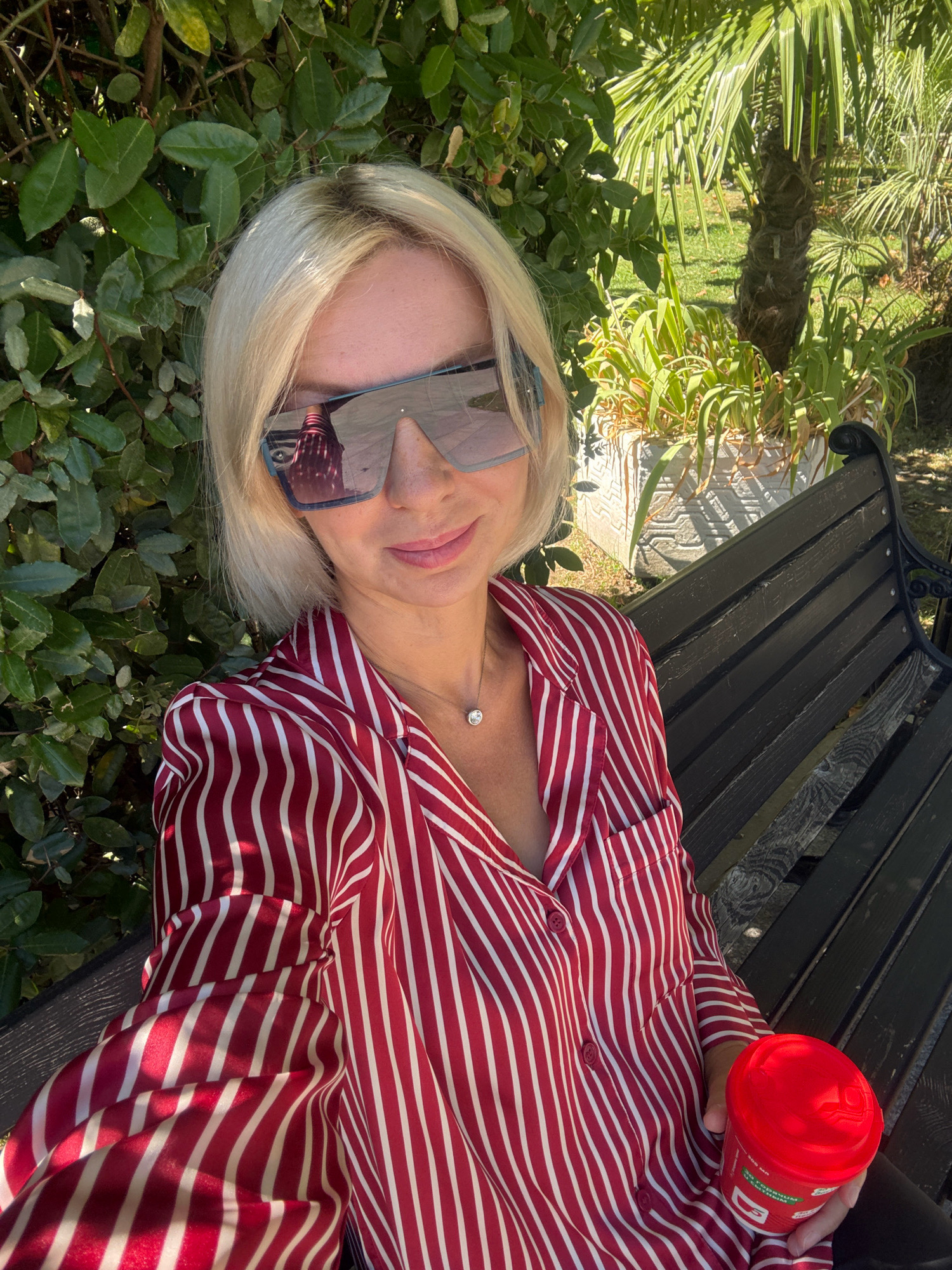 A blonde woman wearing a red and white striped pajama top and large sunglasses is sitting on a dark wooden park bench outdoors. She is holding a red cup in her right hand. The background features lush green foliage and palm trees, indicative of a park setting. The sun is shining, casting shadows. The scene appears to be in a warm climate, consistent with Sochi, Russia.