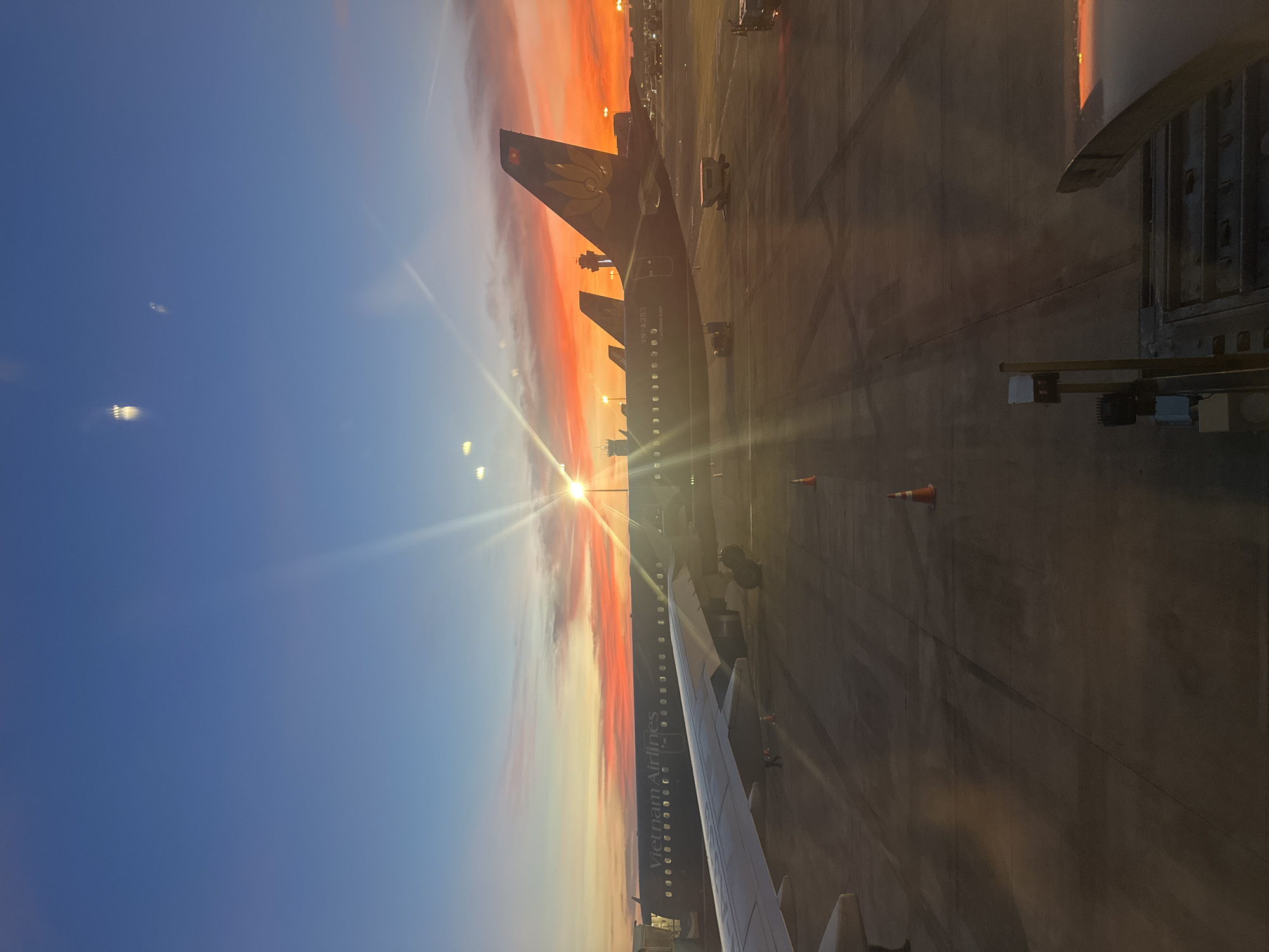 The image captures a vibrant sunset over an airport tarmac in Thuận An, Vietnam. The sky is a dramatic blend of deep blue, fading into hues of orange and red, with the sun's bright rays piercing through, creating lens flare. Several Vietnam Airlines airplanes are parked on the tarmac. The closest aircraft, partially in the foreground, has its wing visible. The fuselage of another aircraft bears the "Vietnam Airlines" logo. In the background, a control tower and other airport structures are silhouetted against the fiery sunset. The tarmac itself is marked with tire tracks and dotted with orange traffic cones and various ground service vehicles. The overall scene is one of a busy airport at the end of the day, bathed in the warm, colorful light of the setting sun. There are no people clearly visible in the frame.