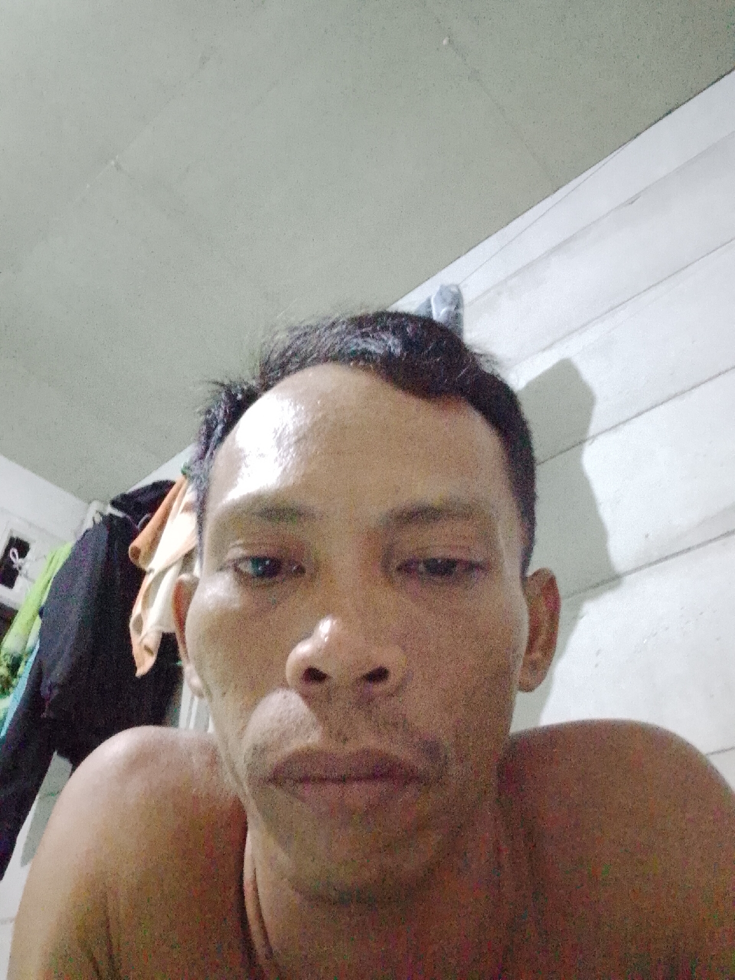 The image shows a close-up selfie of a middle-aged man with dark hair and a bare upper body. His skin appears damp or oily. The background consists of a white wall with horizontal planks and a ceiling with a mottled texture. In the upper left corner, there are out-of-focus clothes hanging. The location is Bukit Ketok, Indonesia.