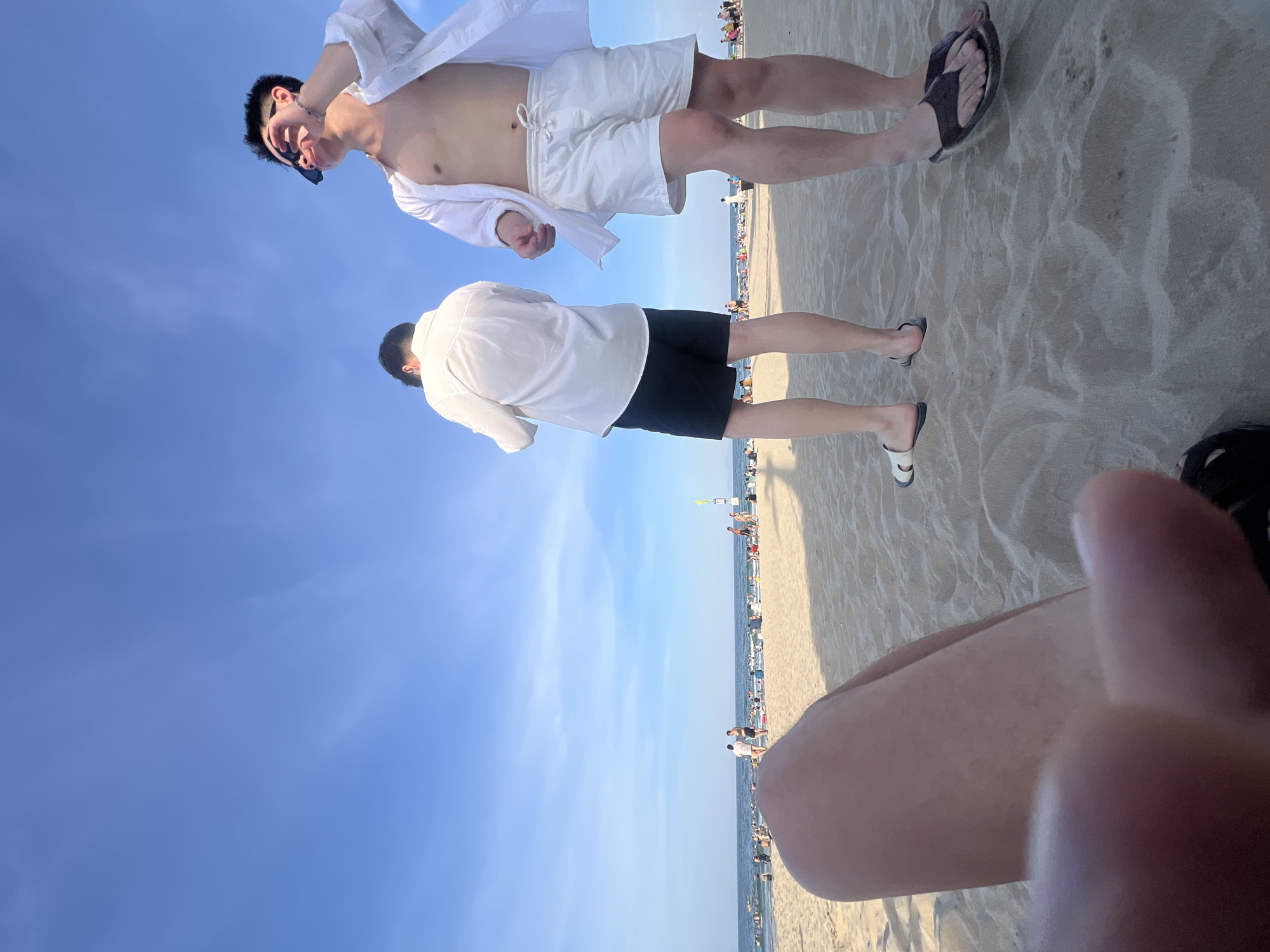 The image shows a group of people on a sunny beach with light blue skies. The foreground is dominated by the blurred legs and hand of a person taking the photo. In the mid-ground, two young men are standing on the sand. The man on the right is wearing white shorts and an unbuttoned white shirt, sunglasses, and black flip-flops. He is looking upwards and to his right, with his hand near his face. The man on the left is wearing black shorts and a white button-up shirt, and is facing away from the camera. Further in the background, a crowded beach stretches along the coast, with the ocean visible beyond. People are seen swimming and relaxing on the shore. The overall setting is consistent with a popular beach destination in Đà Nẵng, Vietnam, known for its sandy coastline and vibrant tourist activity.