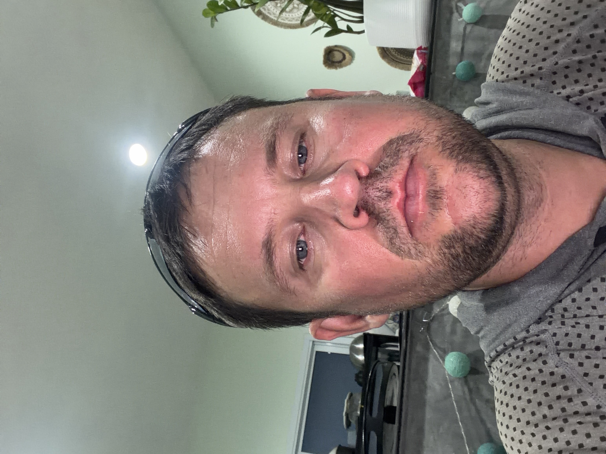 A close-up, eye-level, selfie-style, medium shot shows a Caucasian man in his late 30s or early 40s with fair skin, short brown hair, and a light beard, looking directly at the camera. He is wearing a grey hooded sweatshirt with a subtle patterned design. He has sunglasses pushed up on his forehead. His face is flushed, possibly from heat or exertion, and his skin appears slightly sweaty. His eyes are blue and he has a neutral expression.
The background suggests an indoor setting, possibly a kitchen or living area in a home. To the left, a window with a dark frame is visible, and on a counter behind him, there are what appear to be kitchen appliances or decorative bowls. A string of teal and white spherical decorations hangs across the background, suggesting a festive or decorative element. To the right, a potted green plant with large leaves is visible, along with some woven decorative items on the wall. The lighting is bright and somewhat harsh, with a visible overhead light fixture. The overall impression is casual and personal.
The location is identified as Baan Mae Nam, Thailand.