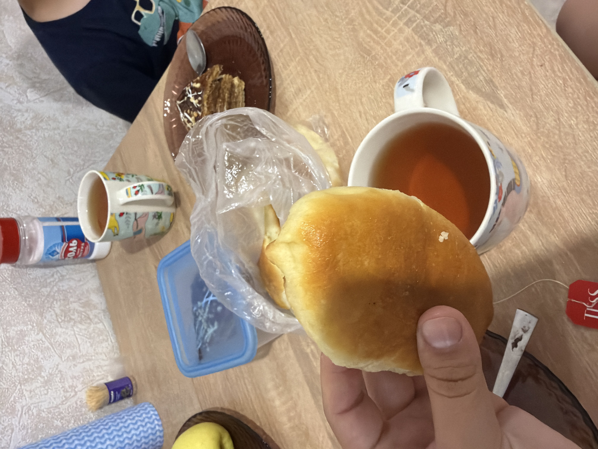 The image shows a close-up view of a wooden table, likely in a home environment in Фролы, Russia, where a meal is being consumed. In the foreground, a person's hand is holding a golden-brown baked bun. To the right of the bun, a white ceramic mug filled with amber-colored tea is visible, with a portion of a teabag tag on the saucer below. In the mid-ground, a clear plastic bag contains more baked goods, and behind that, a blue plastic food container is open. Further back, a slice of layered cake with chocolate frosting and white icing is on a brown plate, with a spoon. To the left of the tea mug, a salt shaker with a red lid and a label reading "СОЛЬ" (salt) is present. A small decorative item, possibly a miniature brush, is to the left of the salt shaker, and a blue and white patterned cloth is partially visible in the upper left corner. In the background, a portion of a child wearing a dark t-shirt with a dinosaur graphic is visible, seated at the table. The overall setting appears to be a casual dining scene.
