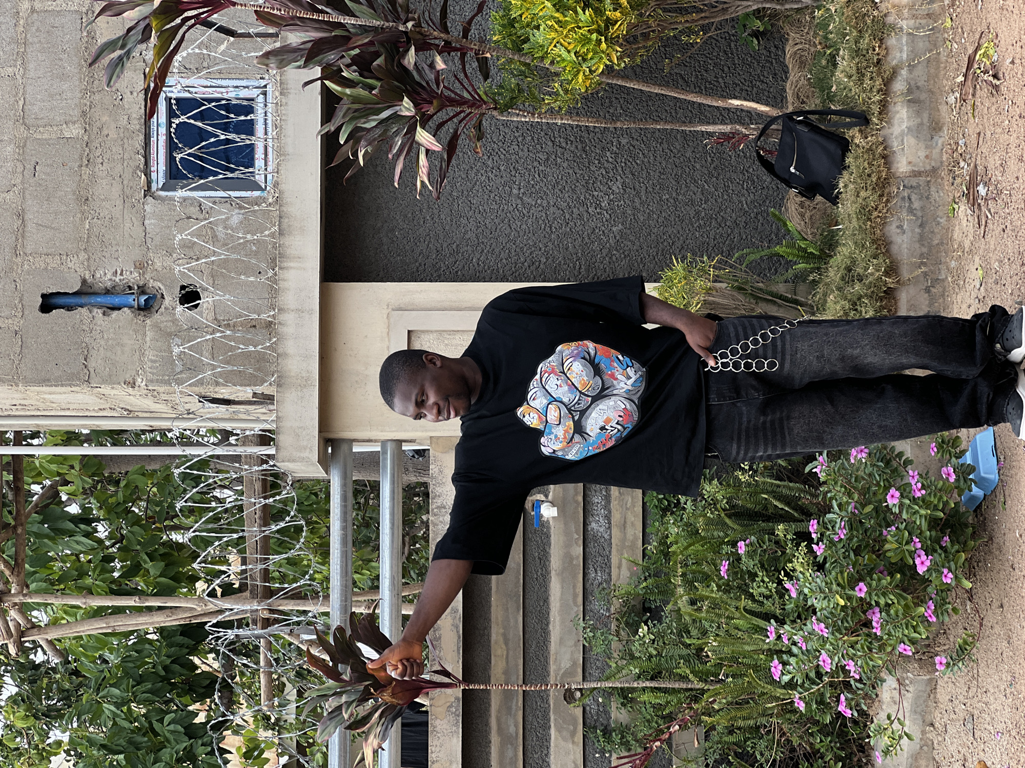 A medium shot depicts a dark-skinned man standing outside a building with barbed wire. He is wearing a black t-shirt with a colorful cartoon graphic, black pants with a silver chain, and sneakers. He is holding the stem of a maroon leafy plant.
The building is constructed from grey concrete blocks with a small window protected by barbed wire. More barbed wire is also visible along the building perimeter. Plants, including flowering ones, surround the building. A black backpack lies on the ground near one of the plants. A set of concrete stairs with metal railings are also visible near the building.
The location is Bukuru, Nigeria, inferred from GPS data.