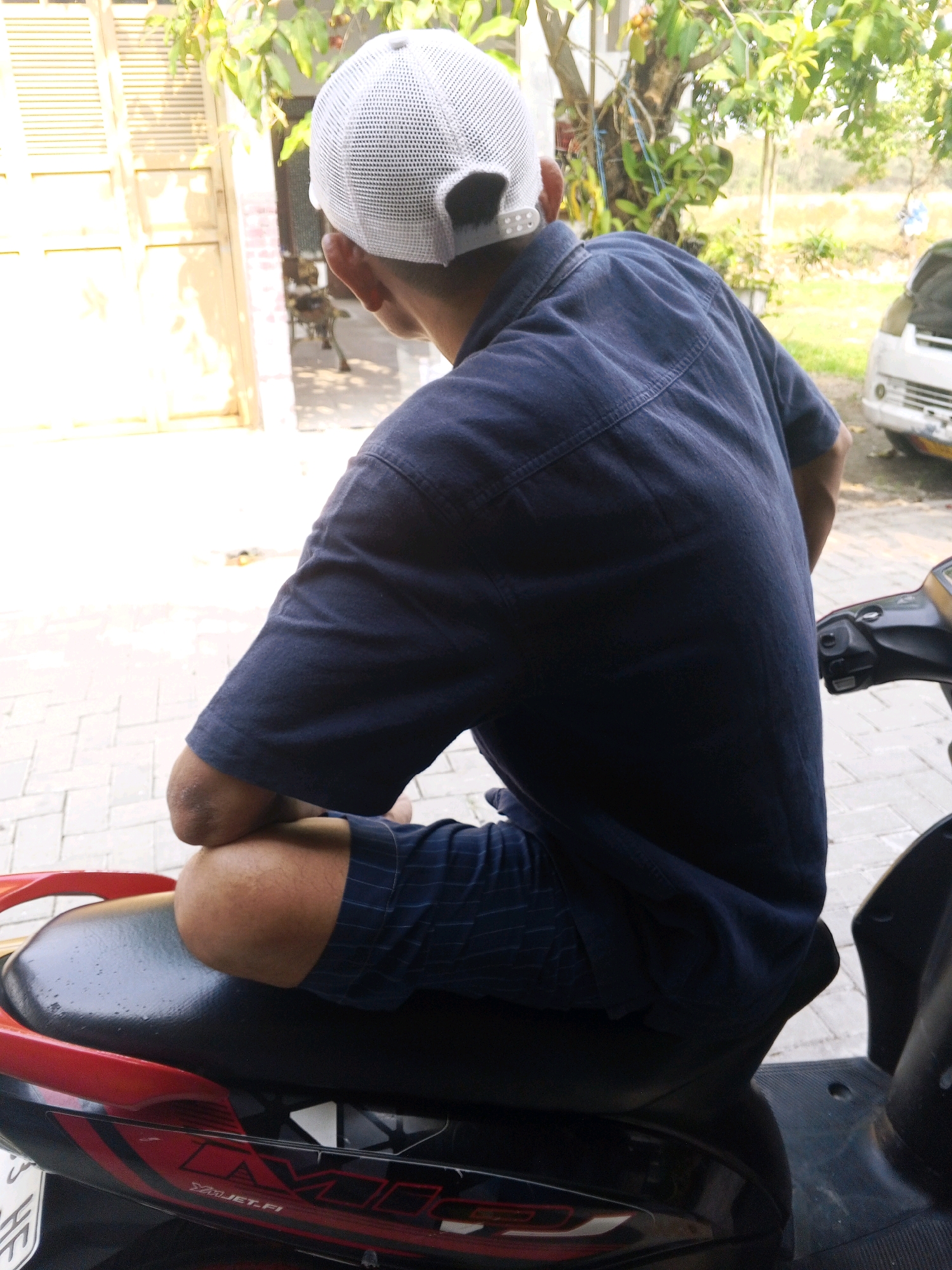 A person wearing a dark blue collared shirt and dark blue checkered shorts sits on a red and black motorcycle. The person is also wearing a white mesh baseball cap. The motorcycle is parked on a paved surface with some sunlight casting shadows. In the background, there is a building with a closed door and shutters, as well as trees and foliage. A white car is visible in the far right of the image. The scene is consistent with a residential or commercial area in Ciruas, Indonesia.