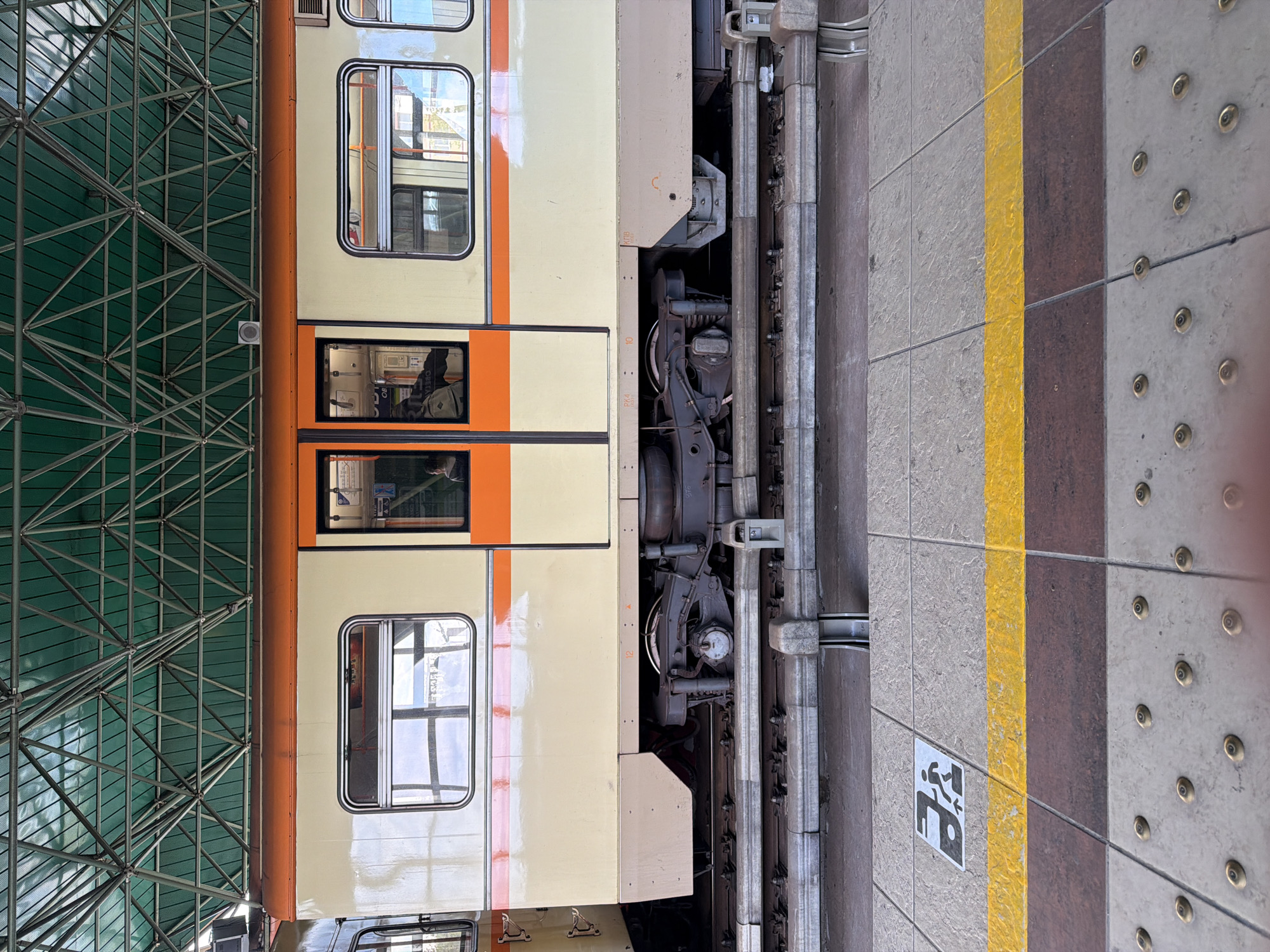A view of a train at a station in Sofia, Bulgaria. The train, cream-colored with orange accents, is next to a platform that is marked with a yellow safety line and a wheelchair symbol. The wheels of the train can be seen along the tracks. The station has a metal structure overhead.