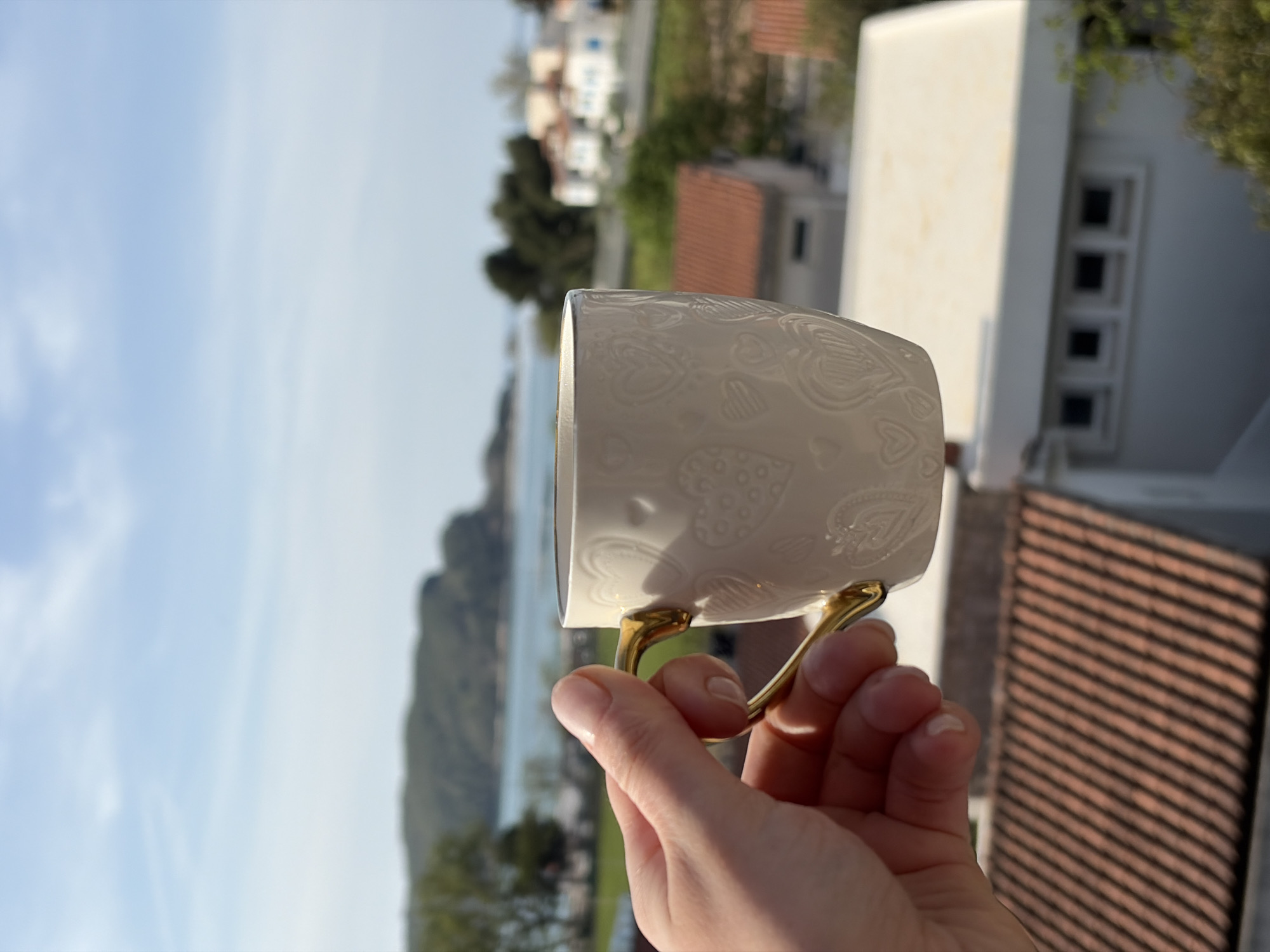 A hand holds a white ceramic mug with a decorative heart pattern and gold-colored handles. The background features a partial view of a city, likely Budva, Montenegro. The sea is visible in the distance, with tree-covered hills bordering it. In the blurred foreground, there is a reddish-brown tiled roof and a white building. The sky is blue with a few clouds. The image is rotated clockwise.