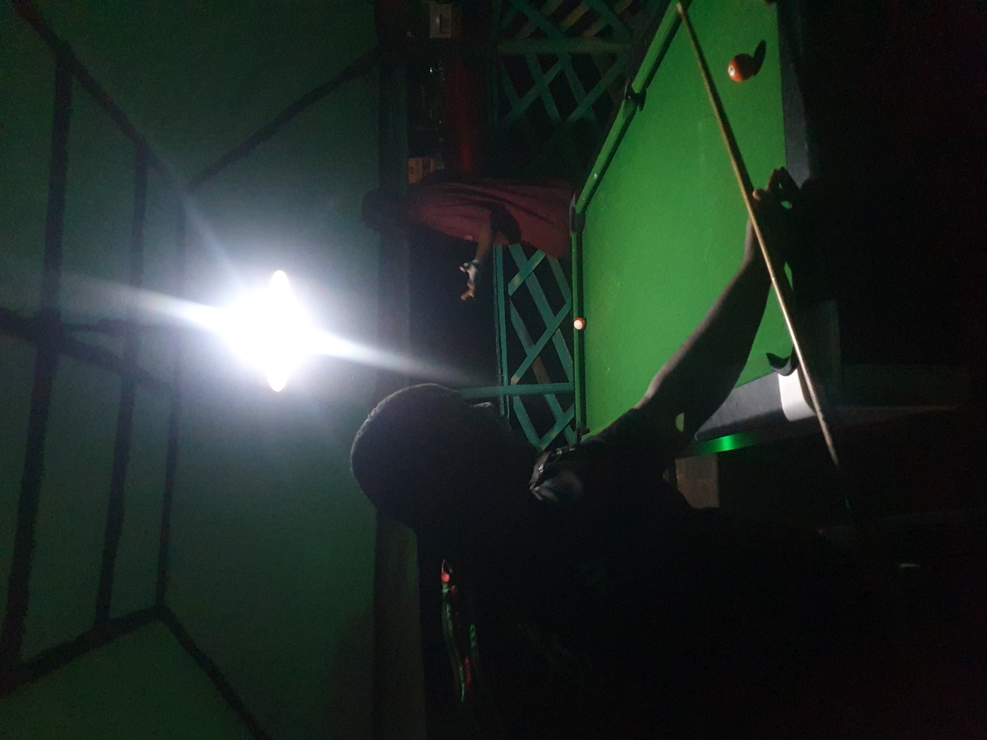 In Lokoja, Nigeria, a dimly lit, open-air pavilion features a green billiard table. Two men are present: one in the foreground, silhouetted against a bright overhead light, is positioned to strike a billiard ball with a cue stick. Another man, wearing a red shirt, stands in the background, gesturing with his hands. The pavilion's green, angled roof is visible, along with a latticework fence at the back, suggesting an outdoor recreational area. The lighting is harsh, creating strong contrasts and obscuring details in the shadows.