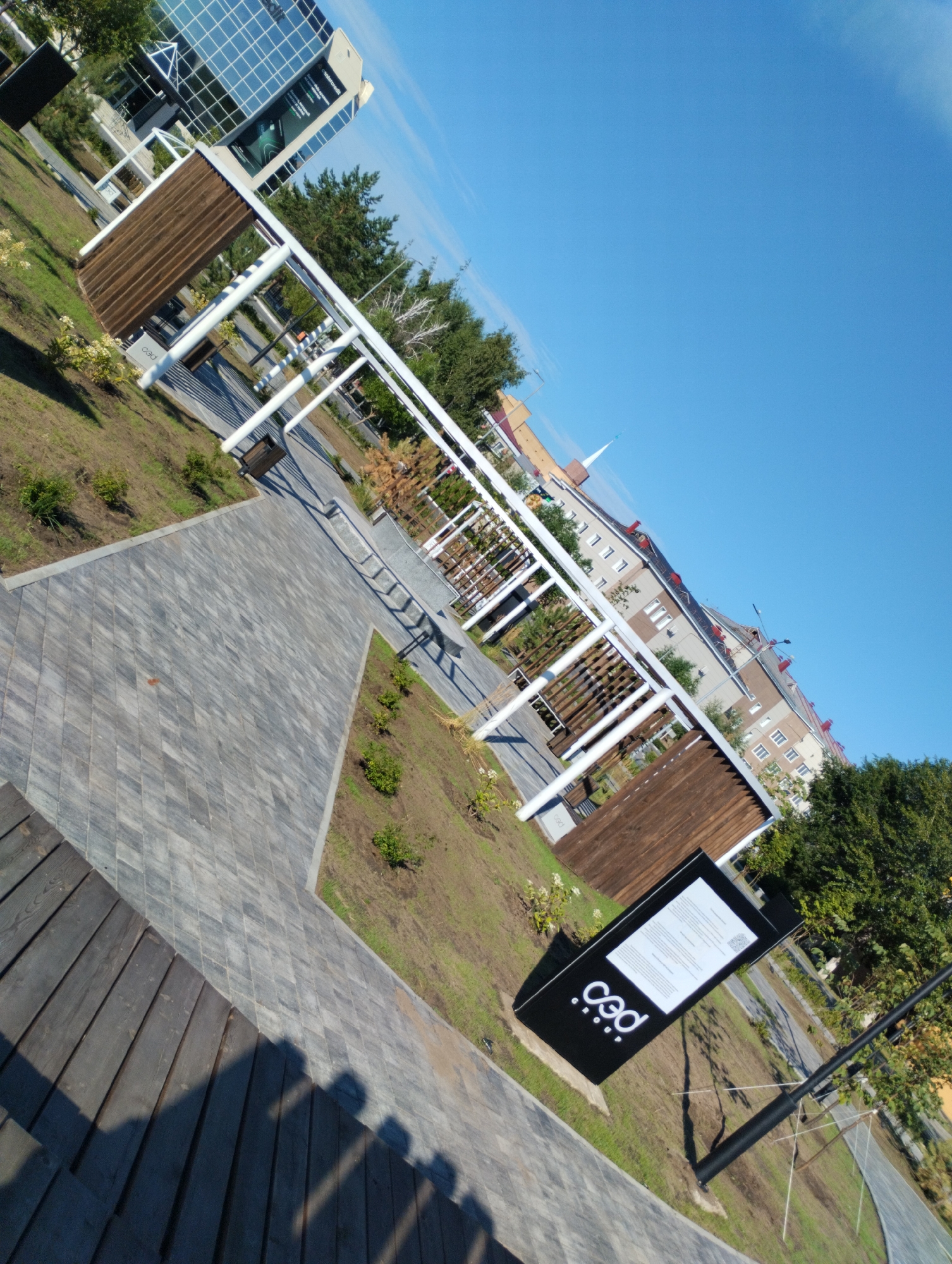 The image shows an outdoor public space with a modern design, likely in the city of Kostanay, Kazakhstan. In the foreground, a dark wooden bench is visible, casting shadows on the paved walkway. The walkway is made of grey diamond-shaped pavers. Beyond the walkway, there is a grassy area with landscaping and several modern white pergolas with wooden slatted infills. A black information stand with a white screen and a logo is positioned near the walkway. In the background, a multi-story building with a glass facade and several residential buildings with red roofs are visible. The sky is clear and blue. The image is taken at an angle.