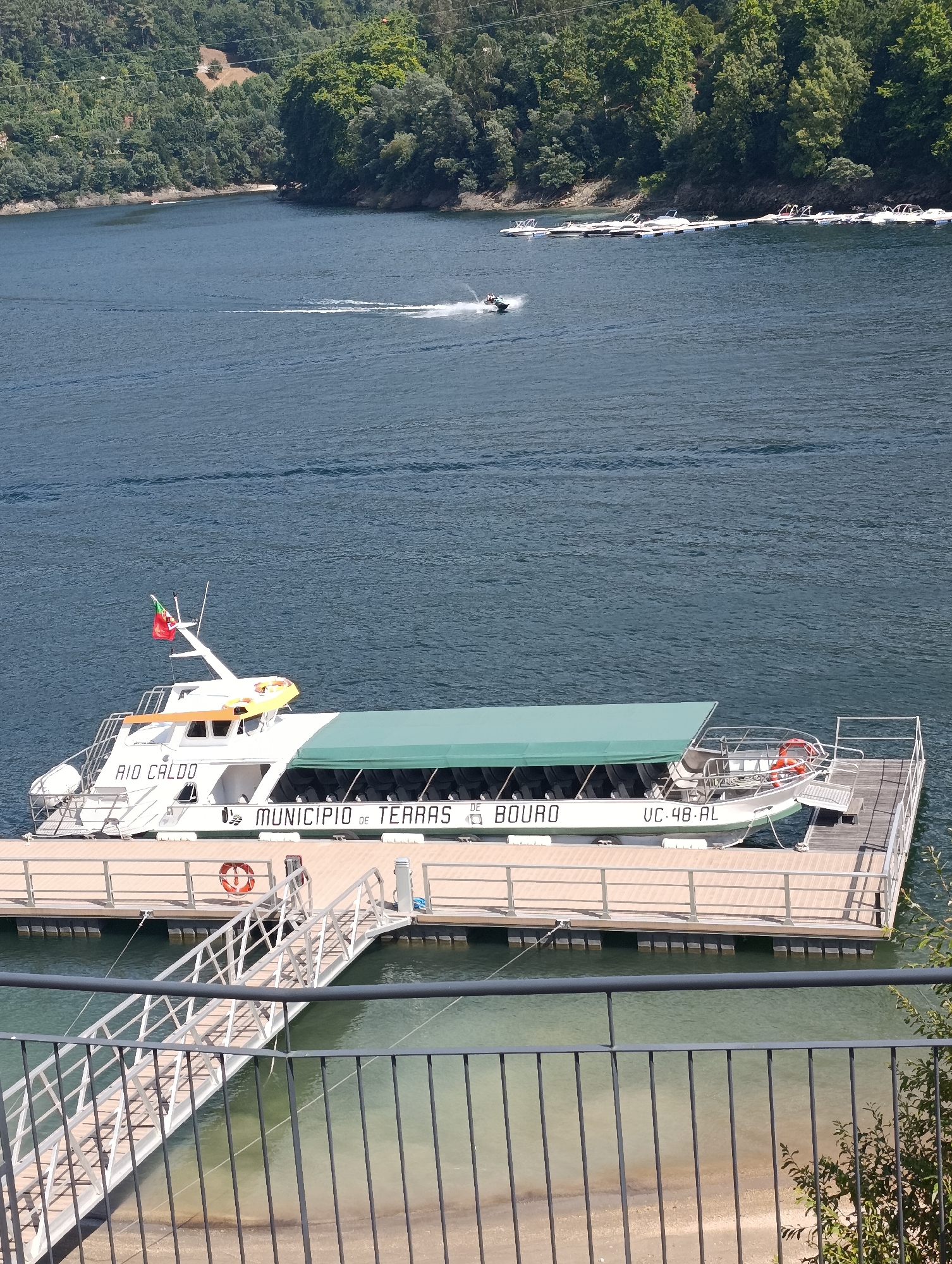 The image captures a view of a large white passenger boat docked at a wooden pier on a body of water. The boat has a green canopy covering the seating area and displays the text "RIO CALDO" and "MUNICIPIO DE TERRAS DE BOURO" along its hull, along with the registration number "UC.48.AL". A Portuguese flag is visible on the boat's mast. The pier is equipped with railings and a ramp leading to the shore. In the background, a lush green, tree-covered hillside slopes down to the water. Several smaller boats are moored along a floating dock in the distance, and a jet ski can be seen creating a wake on the water. The scene appears to be in Terras de Bouro, Portugal, given the boat's inscription.