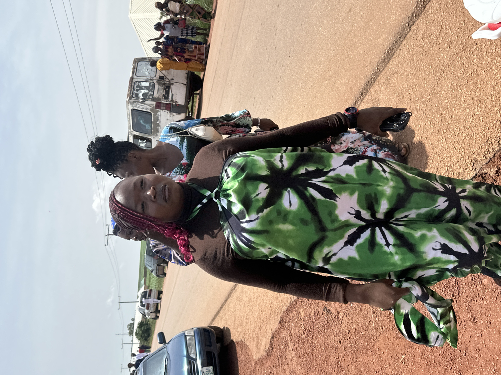 In Durumi, Nigeria, a group of people are gathered on the side of a paved road. The focal point is a woman in the foreground, wearing a brown long-sleeved shirt and a green and white tie-dye dress. She has bright pink braids and is holding a dark object, possibly a phone, in her right hand. Behind her, other individuals are visible, including a woman with her hair in a bun and another wearing a colorful patterned dress. Several vehicles are present, including a blue car on the left and a white van further down the road. The environment appears to be a rural or semi-rural setting, with a dirt shoulder on the left and a dusty road stretching ahead. The sky is overcast.