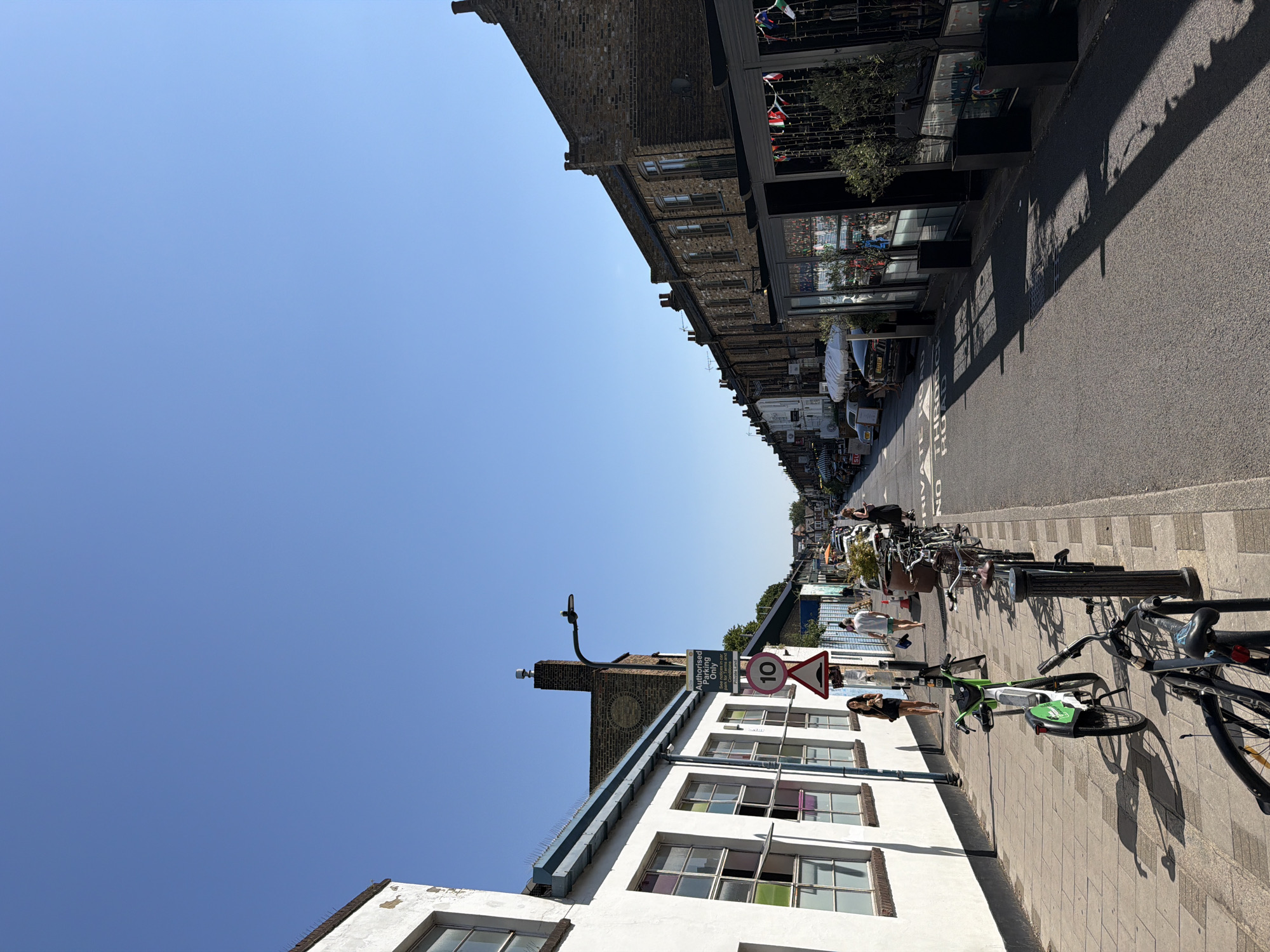The image depicts a street scene in London, United Kingdom, on a clear, sunny day. The street is lined with buildings on both sides. On the left, a white building with large, multi-paned windows, some of which have colored glass, runs along the sidewalk. A sign indicating "Authorised Parking Only" and a speed limit sign for "10" are visible. Several bicycles are parked along the sidewalk, including a bright green shared bike.
On the right side of the street, a row of brick buildings with dark-windowed storefronts and awnings stretches into the distance. These buildings appear to house businesses, possibly restaurants or shops, as suggested by the tables and chairs visible outside and decorative flags. A street lamp extends over the road from the left side. The road itself is asphalt and appears to be a public thoroughfare, with some parked cars visible further down. The sky is a clear, bright blue. A few people are walking on the sidewalk on the left side of the street. The overall impression is of a pleasant, bustling urban environment on a summer day.
