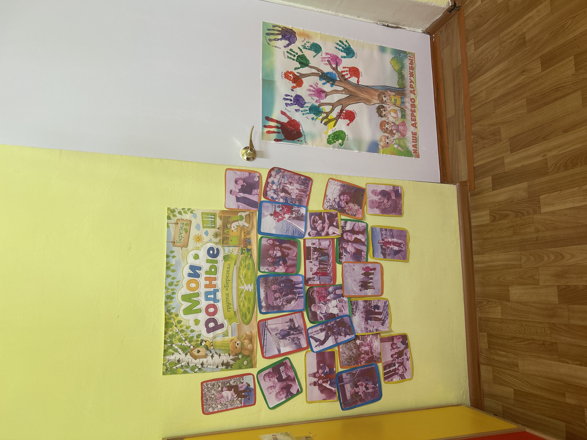 This media file is an indoor shot of a brightly lit wall in what appears to be a classroom or a children's area. The wall is painted in two distinct colors: a pale yellow on the lower half and white on the upper half, with a clean, diagonal division between them.
Prominently displayed on the wall are several pieces of decoration and student work. At the top, on the white section, is a large poster depicting a tree with colorful handprints on its branches, suggesting a children's activity. The poster has text that reads "НАШЕ ДЕРЕВО ДРУЖБЫ!" (Our Friendship Tree!). To the left of this poster, a brass door handle is visible.
Below the large poster, on the yellow section of the wall, is a vertical poster with the title "Мои родные" (My Relatives) in large, colorful letters. This poster also features illustrations of birch trees and a small birdhouse, with the text "группа «Березка»" (group "Berezka") above it.
Scattered around the "Мои родные" poster are numerous smaller, rectangular photographs. These photos appear to be snapshots of groups of people, likely children and adults, in various poses and settings, possibly from events or everyday life. The photographs have colorful borders, primarily blue and yellow.
The floor visible on the right side of the image is made of wooden planks arranged in a typical parquet pattern, suggesting a room with a finished floor. The overall impression is a decorated space for children, filled with personal photos and art projects. The lighting appears to be artificial, with no indicators of natural light or specific time of day. The setting is indoors, likely a school or a kindergarten in Gelendzhik, Russia, given the context.